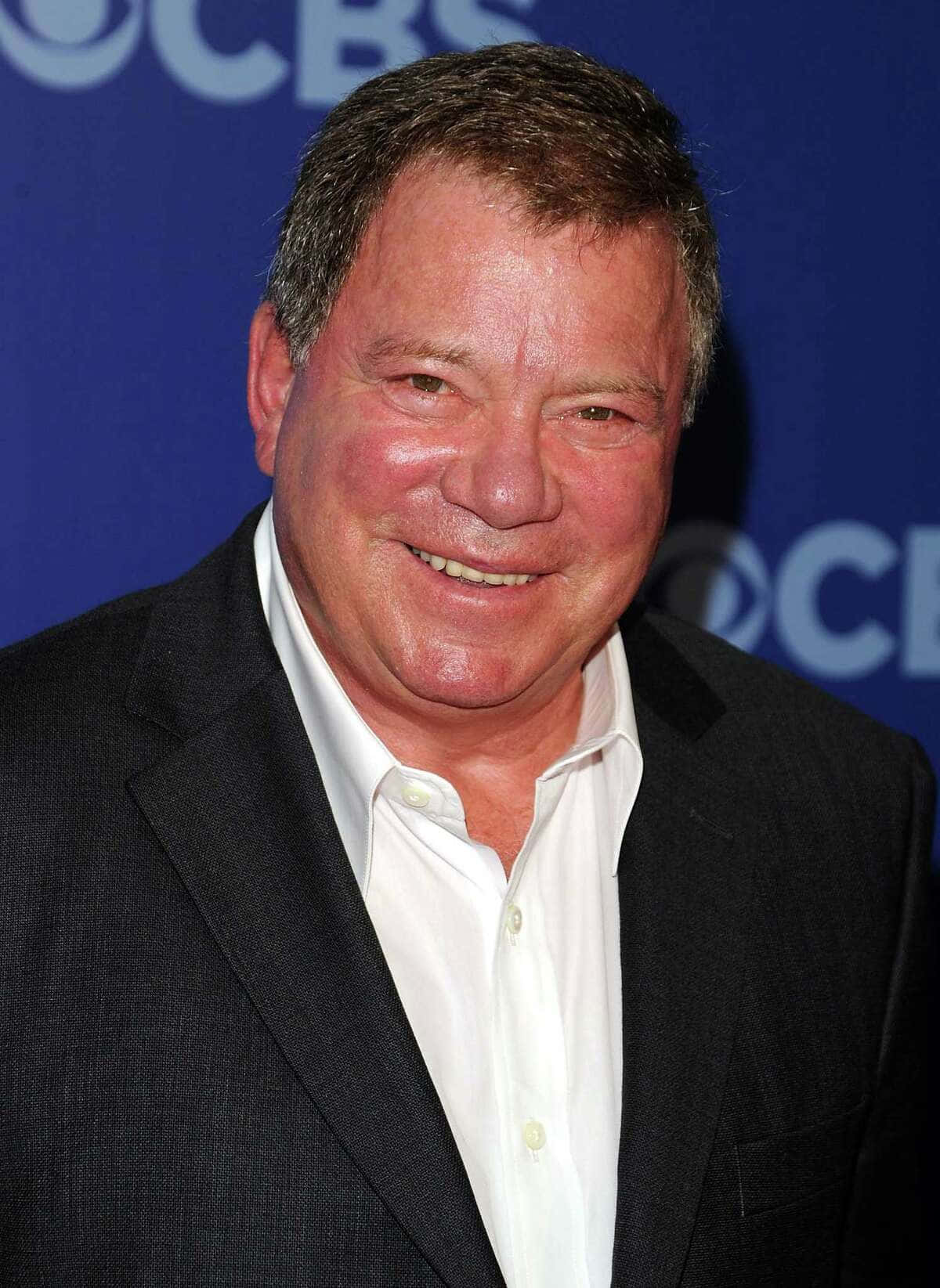 william shatner 桌布 壁紙 — 一位微笑的男子身穿深色西裝和白襯衫，在活動中對著藍色CBS背景擺姿勢。