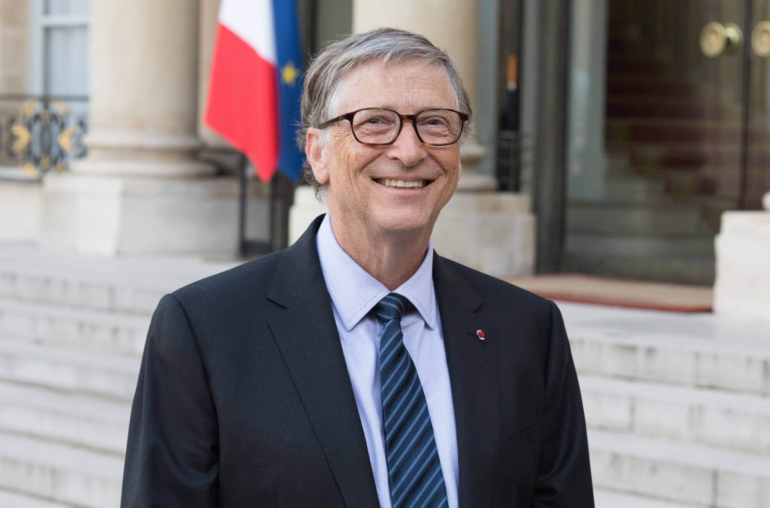 gates 桌布 壁紙 — 一位微笑的男子身穿深色西裝和眼鏡，站在戶外，背景是一座掛有法國國旗的建築物。