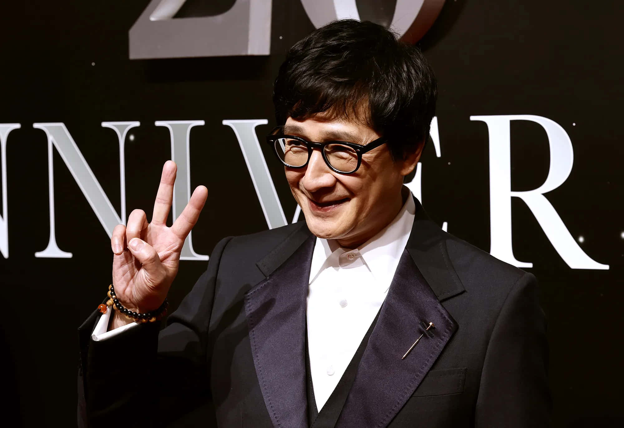 Ke Huy Quan 桌布 壁紙 — 一位微笑的男子身穿深色西裝和眼鏡，在一個帶有「ANNIVERSARY」背景的正式活動中做出和平手勢。