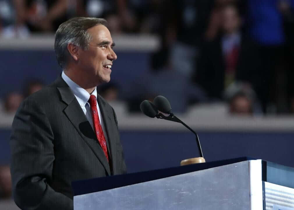 Politics 桌布 壁紙 — 一位微笑的男子身穿深色西裝和紅色領帶，站在講台上，在明亮的舞台燈光下向模糊的觀眾發表演講。