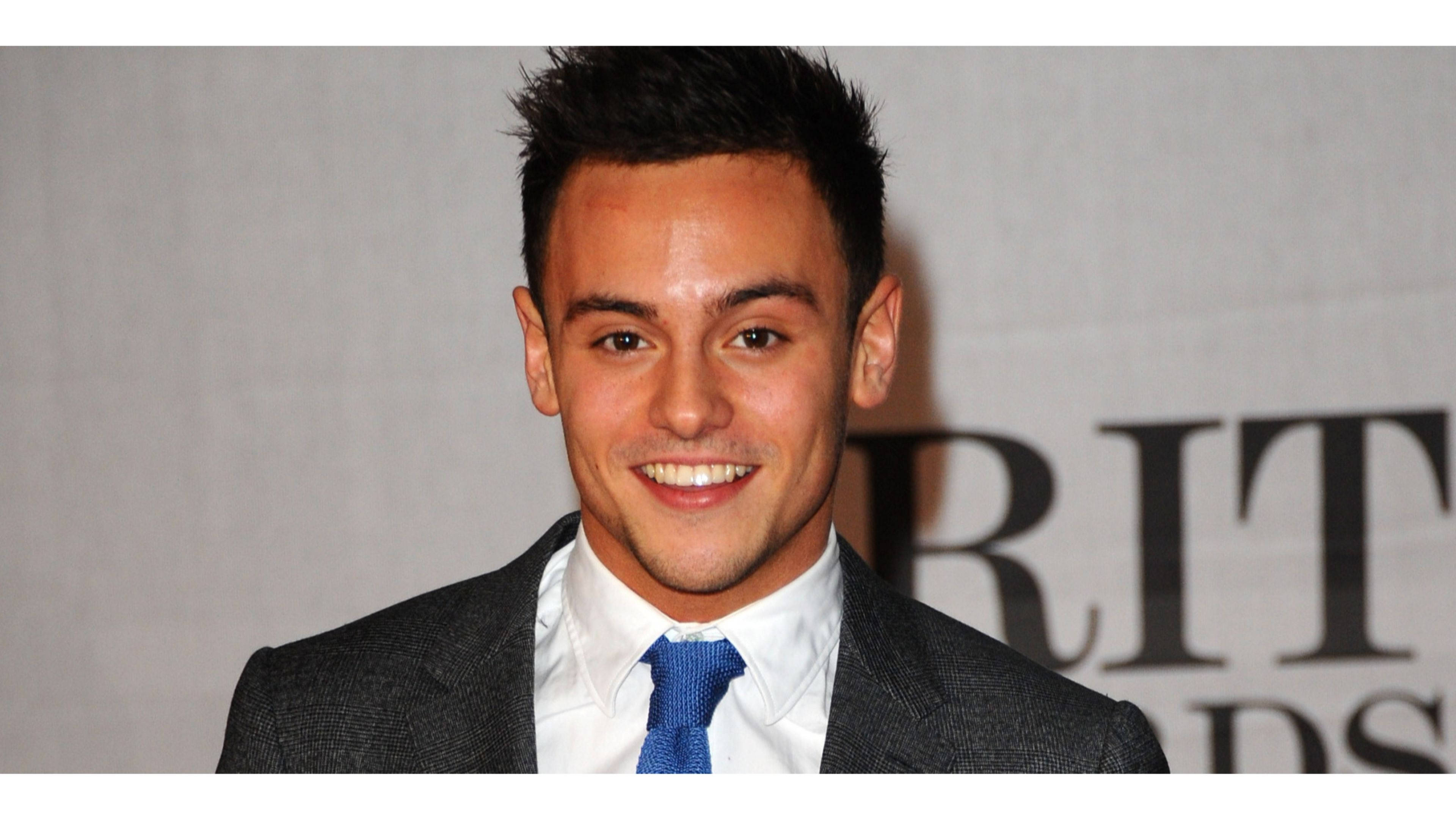 Tom 桌布 壁紙 — 一位微笑的男子身穿深色西裝和藍色領帶，在'BRIT AWARDS'背景的正式活動中自信地擺姿勢。