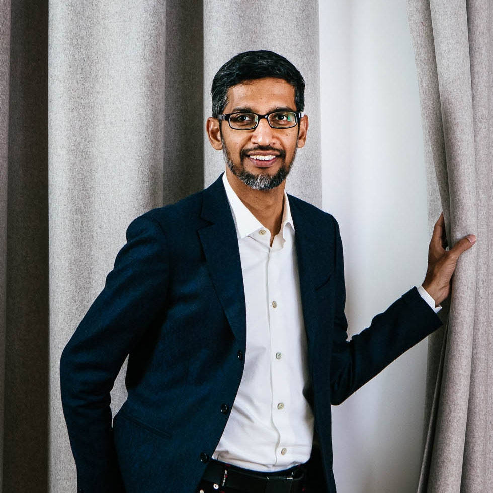 google 桌布 壁紙 — 一位微笑的男子身穿深色西裝外套和眼鏡，自信地站在淺灰色窗簾旁，處於柔和光線的室內環境中。