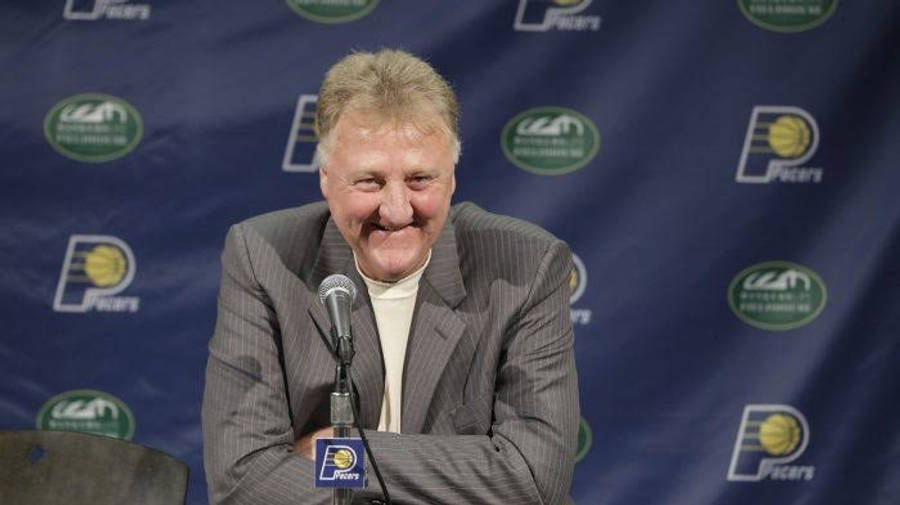NBA 桌布 壁紙 — 一位微笑的男子身穿灰色細條紋西裝，坐在記者會的麥克風前，背景是藍色的印第安納溜馬隊。