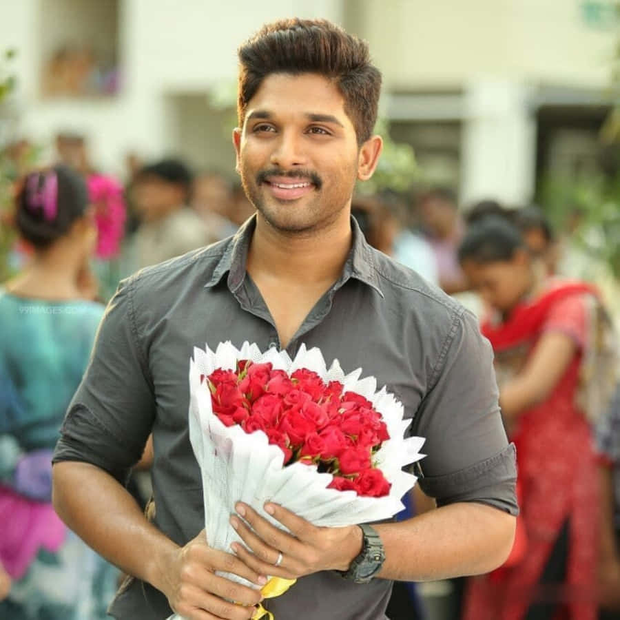 Allu Arjun 桌布 壁紙 — 一位微笑的男子身穿灰色襯衫，手持一束用白紙包裹的鮮豔紅玫瑰，背景是柔和模糊的戶外人群。