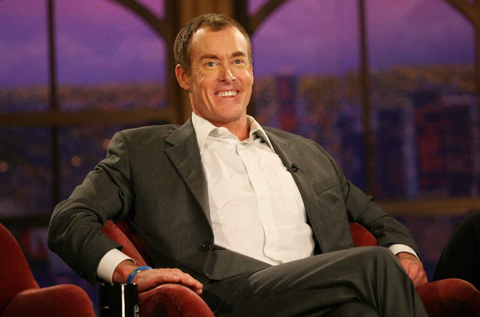 John C. Mcginley 桌布 壁紙 — 一位微笑的男子身穿灰色西裝，放鬆地坐在紅色扶手椅上，背景是模糊的城市天際線的脫口秀場景。