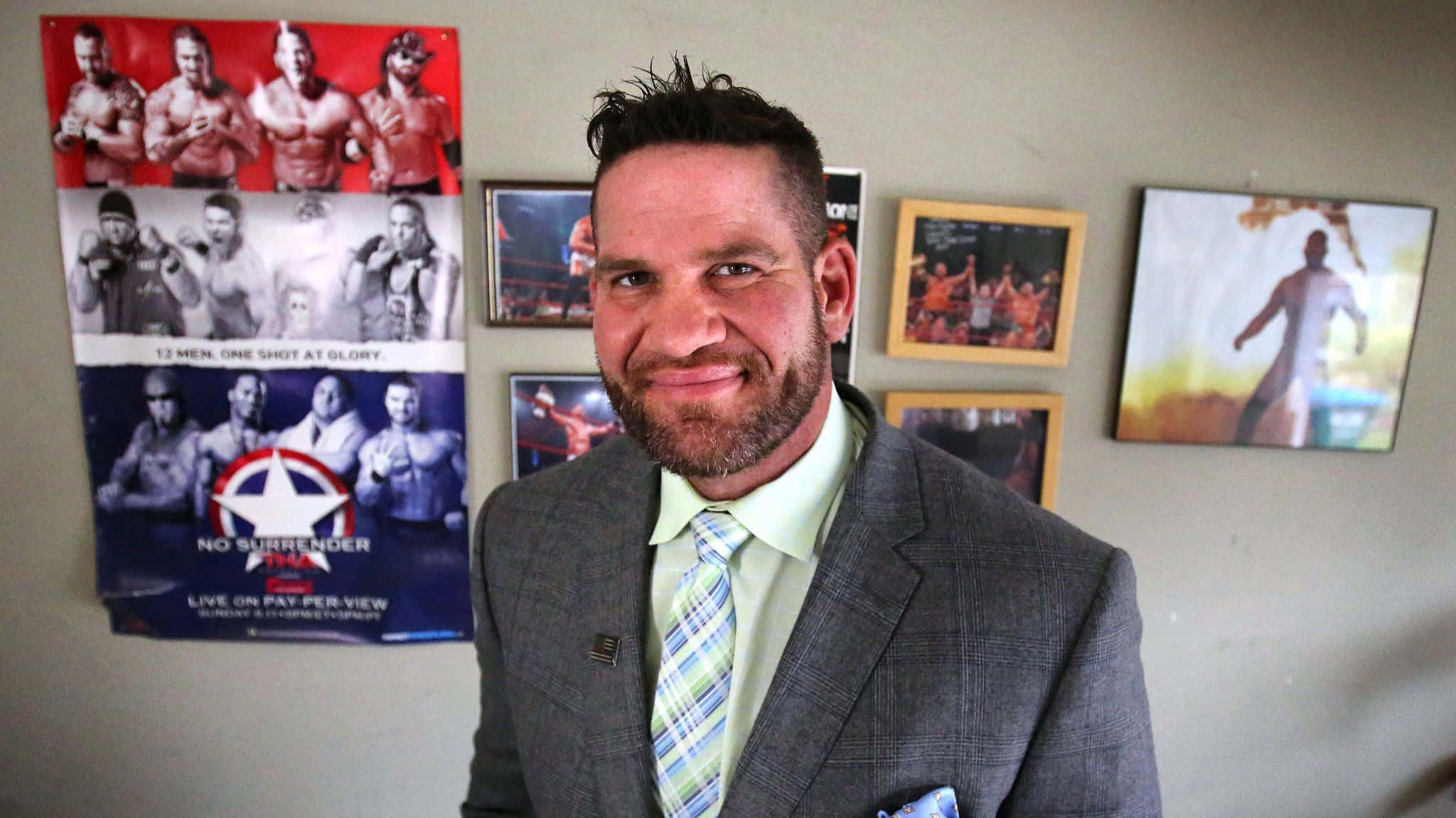 WWE 桌布 壁紙 — 一位微笑的男子身穿灰色西裝和圖案領帶，站在掛滿摔角海報和裱框照片的牆前。