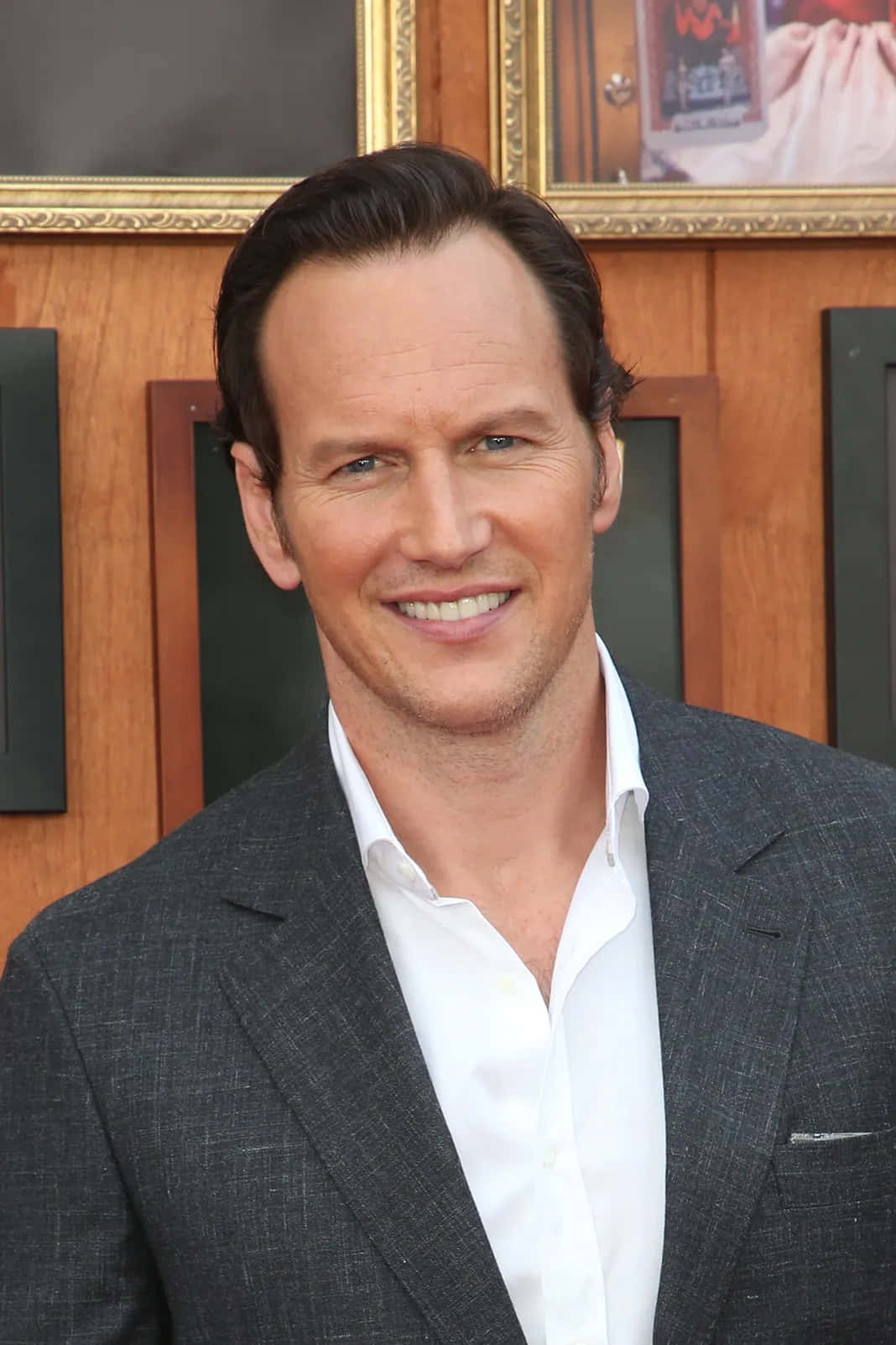 Patrick Wilson 桌布 壁紙 — 一位微笑的男子身穿灰色西裝和白色襯衫，站在掛有裱框圖片的木牆前。