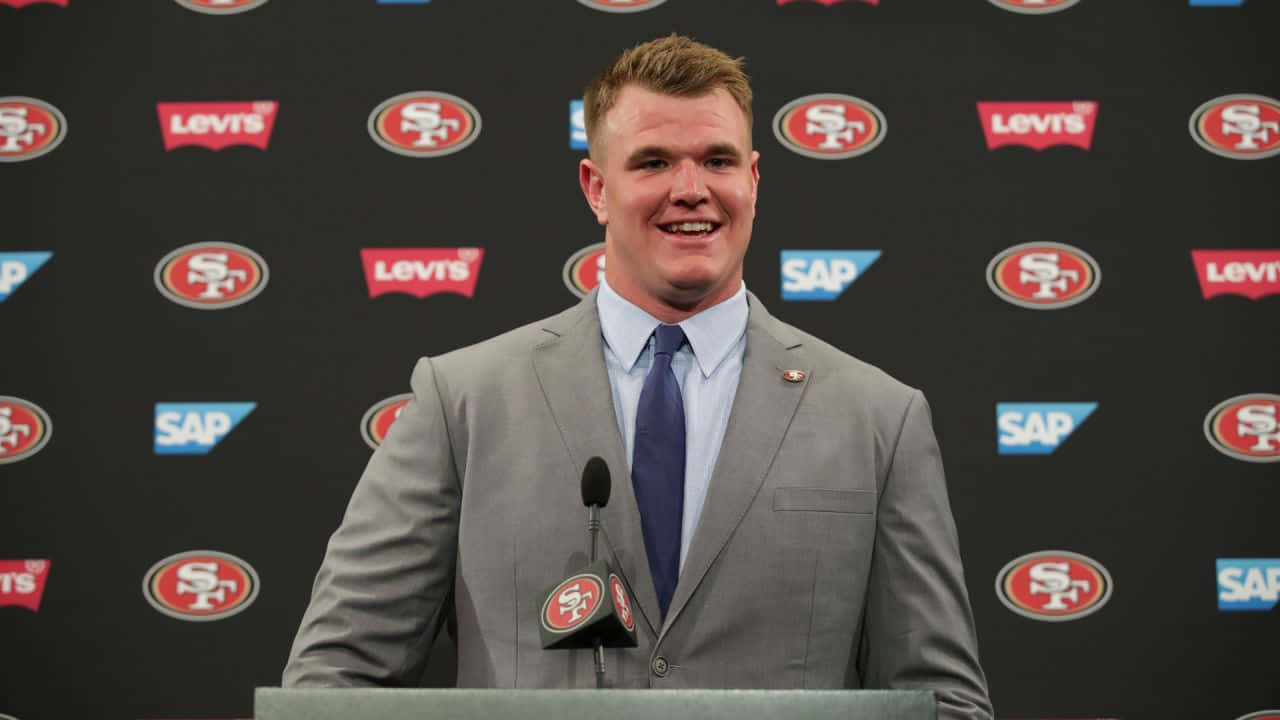 Mike Mcglinchey 桌布 壁紙 — 一位微笑的男子身穿灰色西裝和藍色領帶，站在一個帶有舊金山49人和Levi's標誌的講台上，背景為黑色。