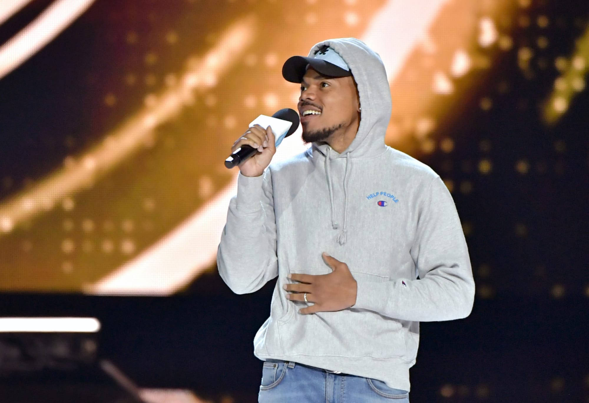 chance 桌布 壁紙 — 一位微笑的男子身穿灰色連帽衫和帽子，在金色光束和散景效果的舞台上對著麥克風唱歌。