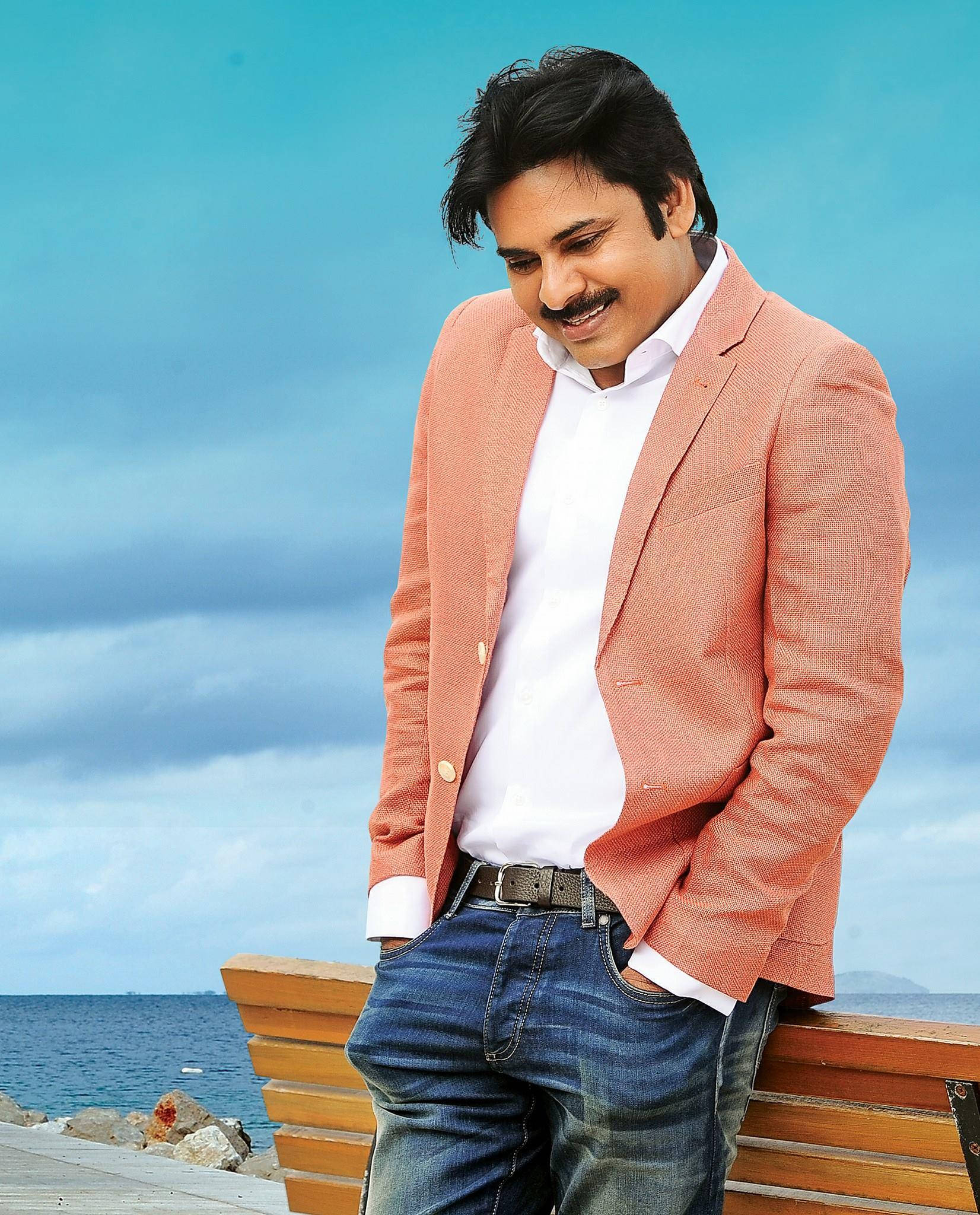 Pawan Kalyan 桌布 壁紙 — 一位微笑的男子身穿珊瑚色西裝外套，在明亮的藍天下隨意地靠在海邊的木長椅上。