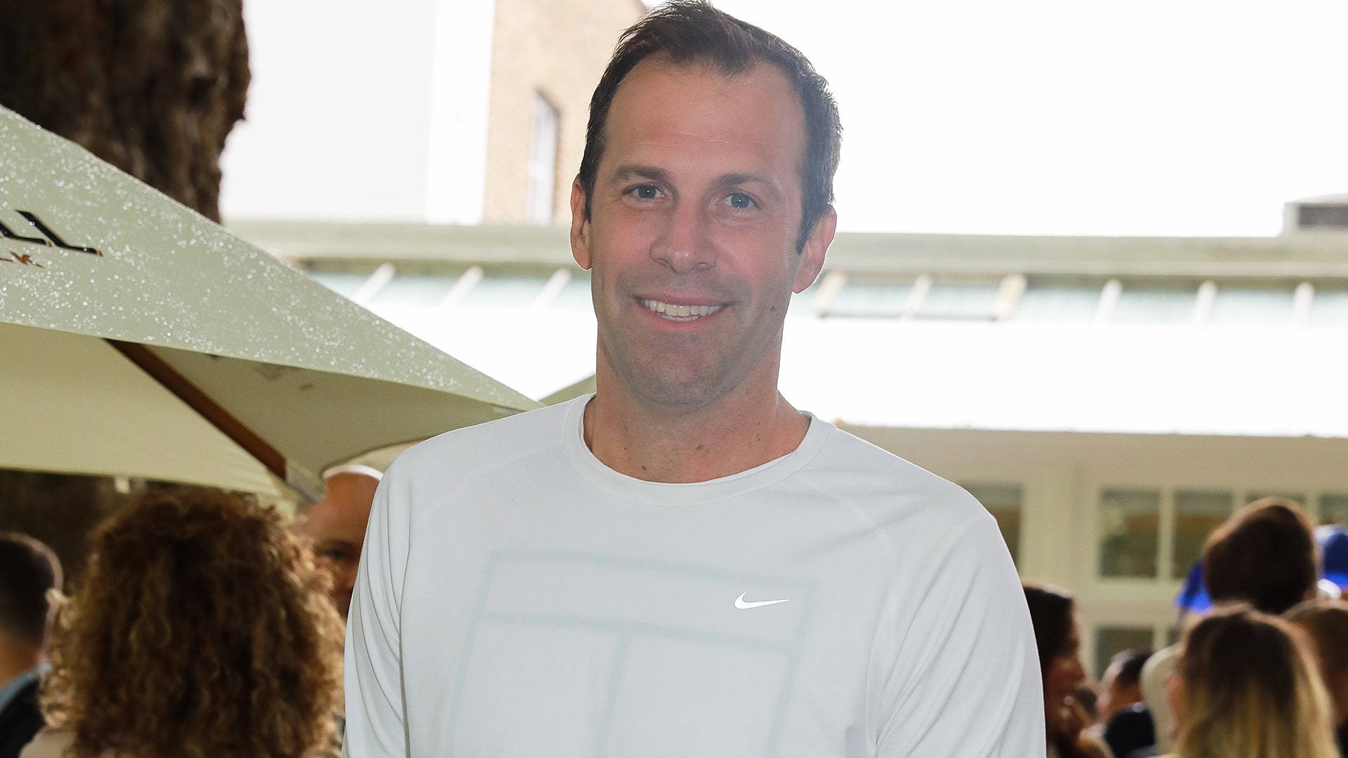運動 桌布 壁紙 — 一位微笑的男子身穿白色Nike網球衫，站在戶外米色遮陽傘下，背景有模糊的人群和建築物。