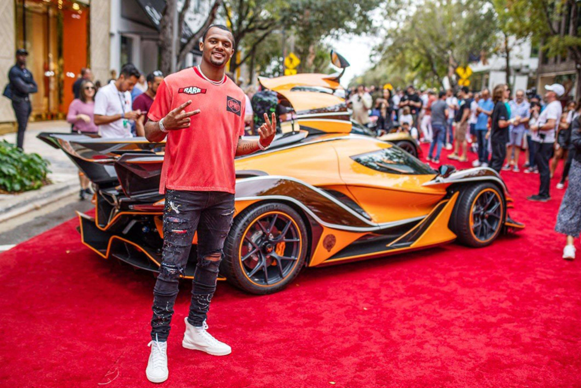 Deshaun Watson 桌布 壁紙 — 一位微笑的男子身穿紅色襯衫，自信地在紅地毯上與一輛引人注目的橙色和黑色超跑合照，周圍有群眾觀看。