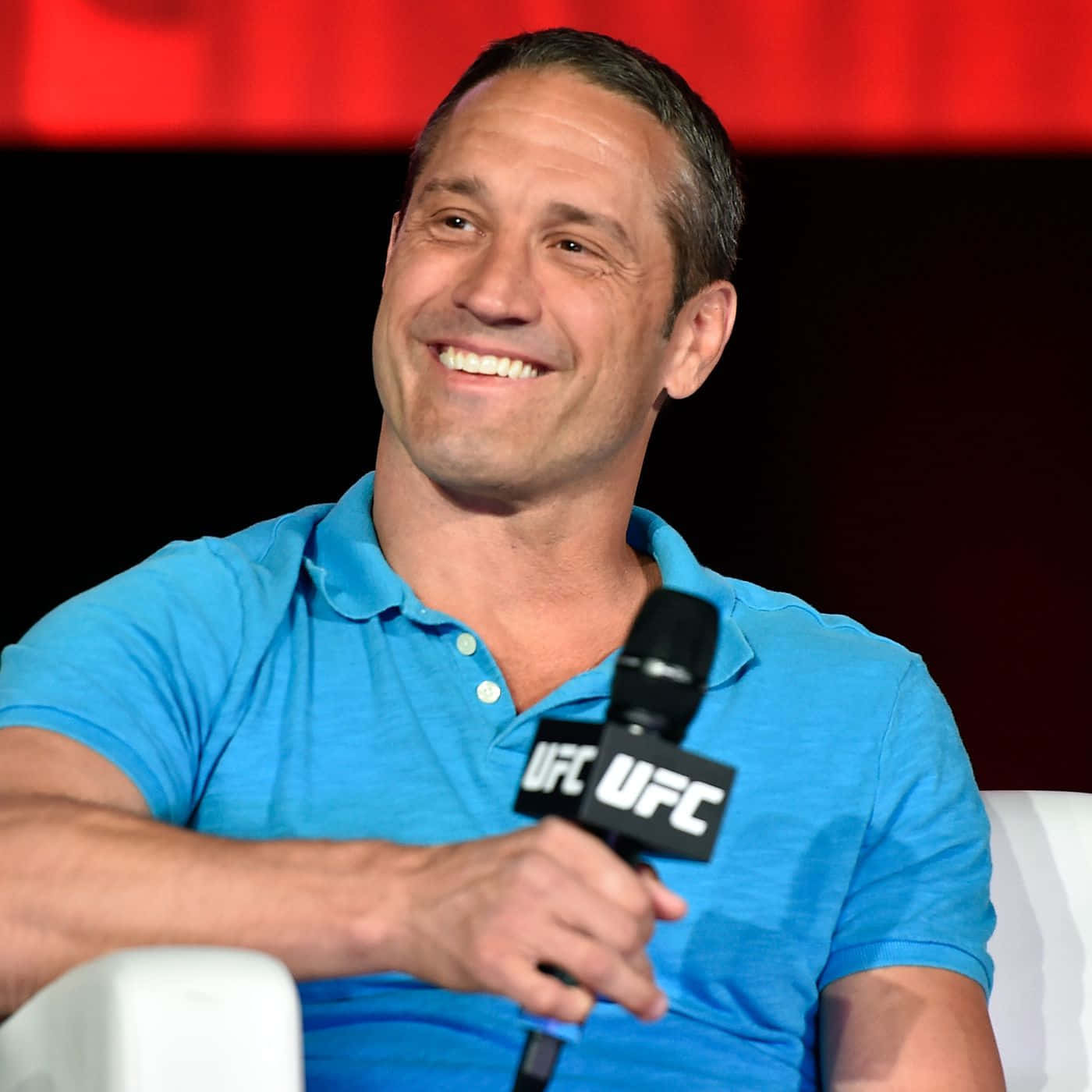UFC 桌布 壁紙 — 一位微笑的男子身穿藍色Polo衫，坐在白色椅子上，手持UFC麥克風，背景為紅色和黑色。