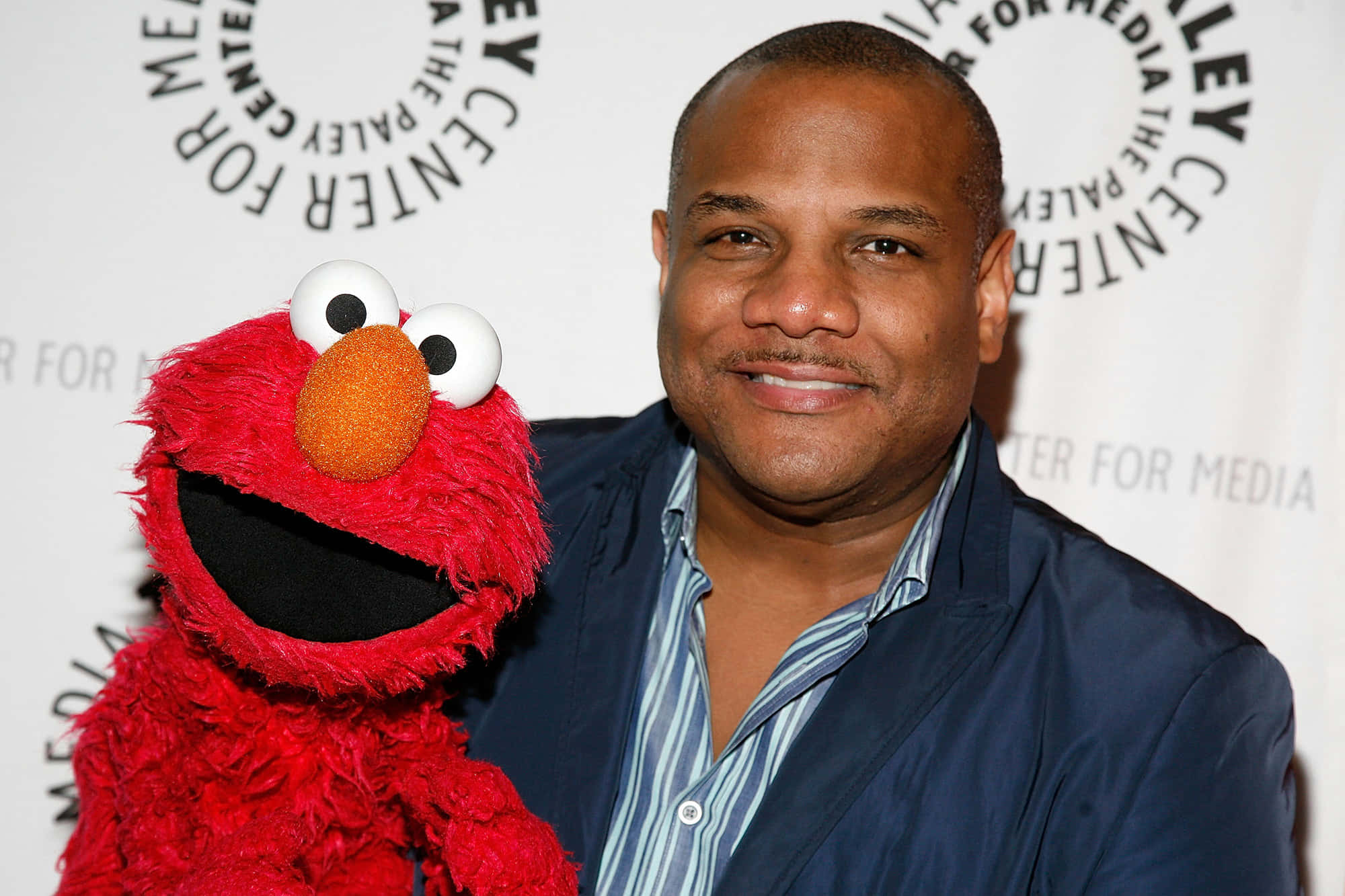 Elmo 桌布 壁紙 — 一位微笑的男子身穿藍色外套，在媒體中心活動中抱著紅色毛絨的Elmo公仔。
