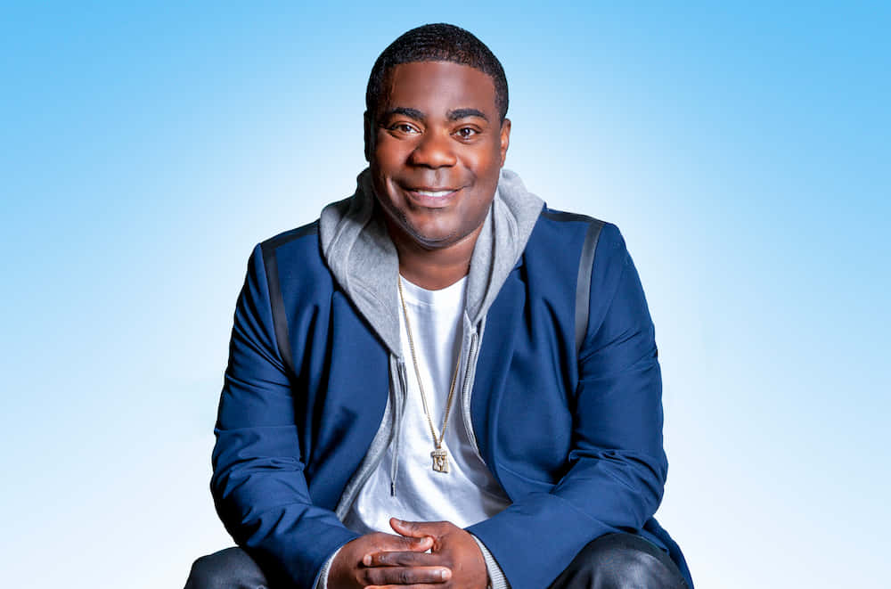 Tracy Morgan 桌布 壁紙 — 一位微笑的男子身穿藍色外套和灰色連帽衫，在柔和的漸變藍色背景前擺姿勢。