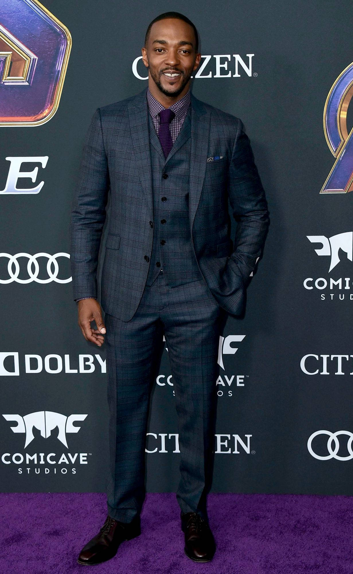 Anthony Mackie 桌布 壁紙 — 一位微笑的男子身穿藍色格紋三件套西裝，自信地站在Marvel活動的紫色地毯上。