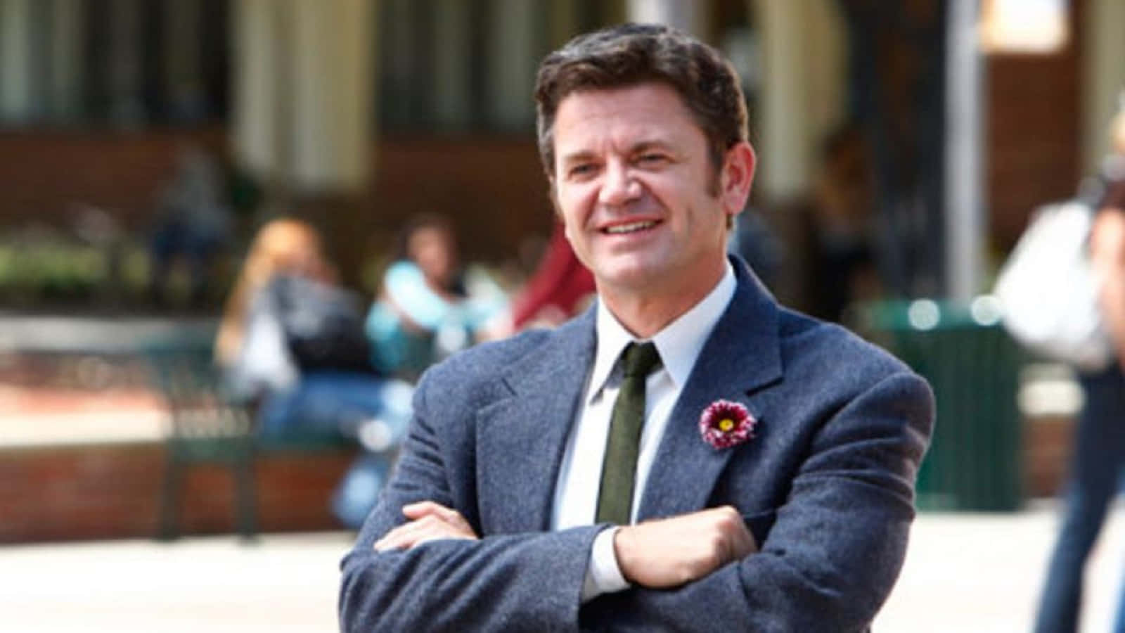 John Michael Higgins 桌布 壁紙 — 一位微笑的男子身穿藍色粗花呢西裝，別著罌粟花胸針，在陽光普照的戶外廣場自信地交叉雙臂站立。