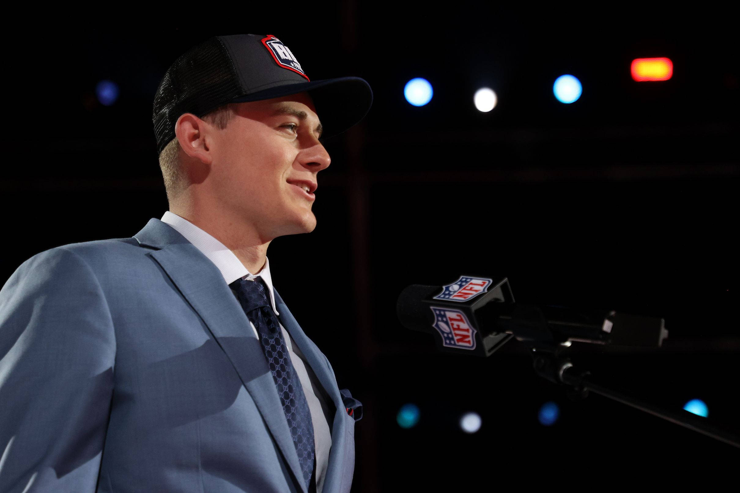 New England Patriots 桌布 壁紙 — 一位微笑的男子身穿藍色西裝和NFL帽子，在舞台燈光下對著麥克風說話。
