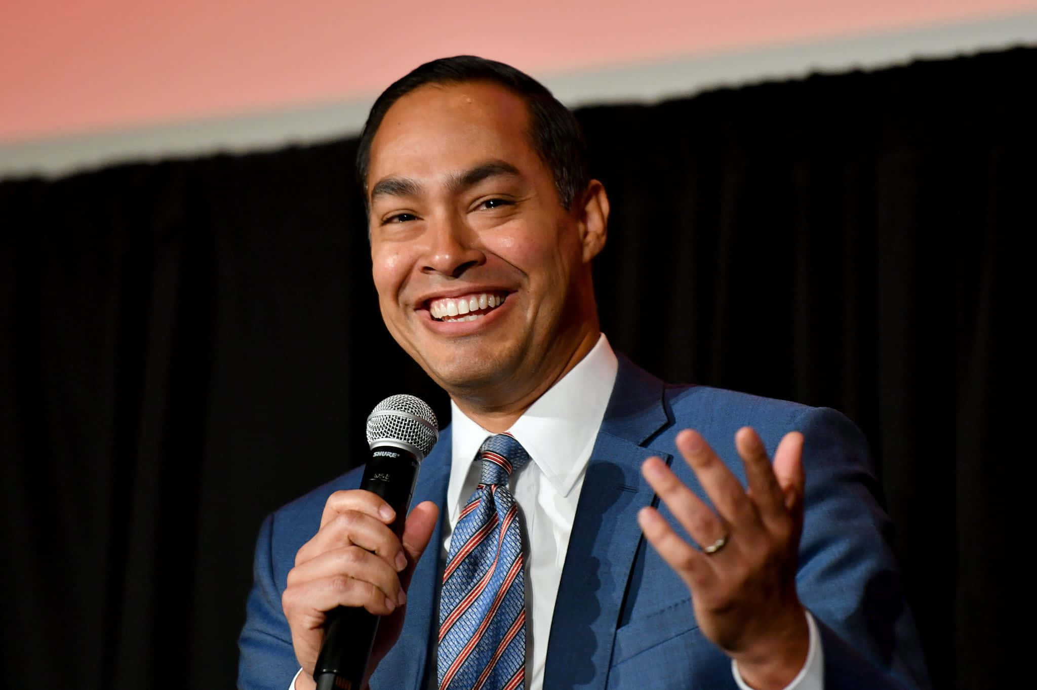 Celebrity 桌布 壁紙 — 一位微笑的男子身穿藍色西裝和圖案領帶，手持麥克風，在黑色窗簾背景下熱情地做手勢。