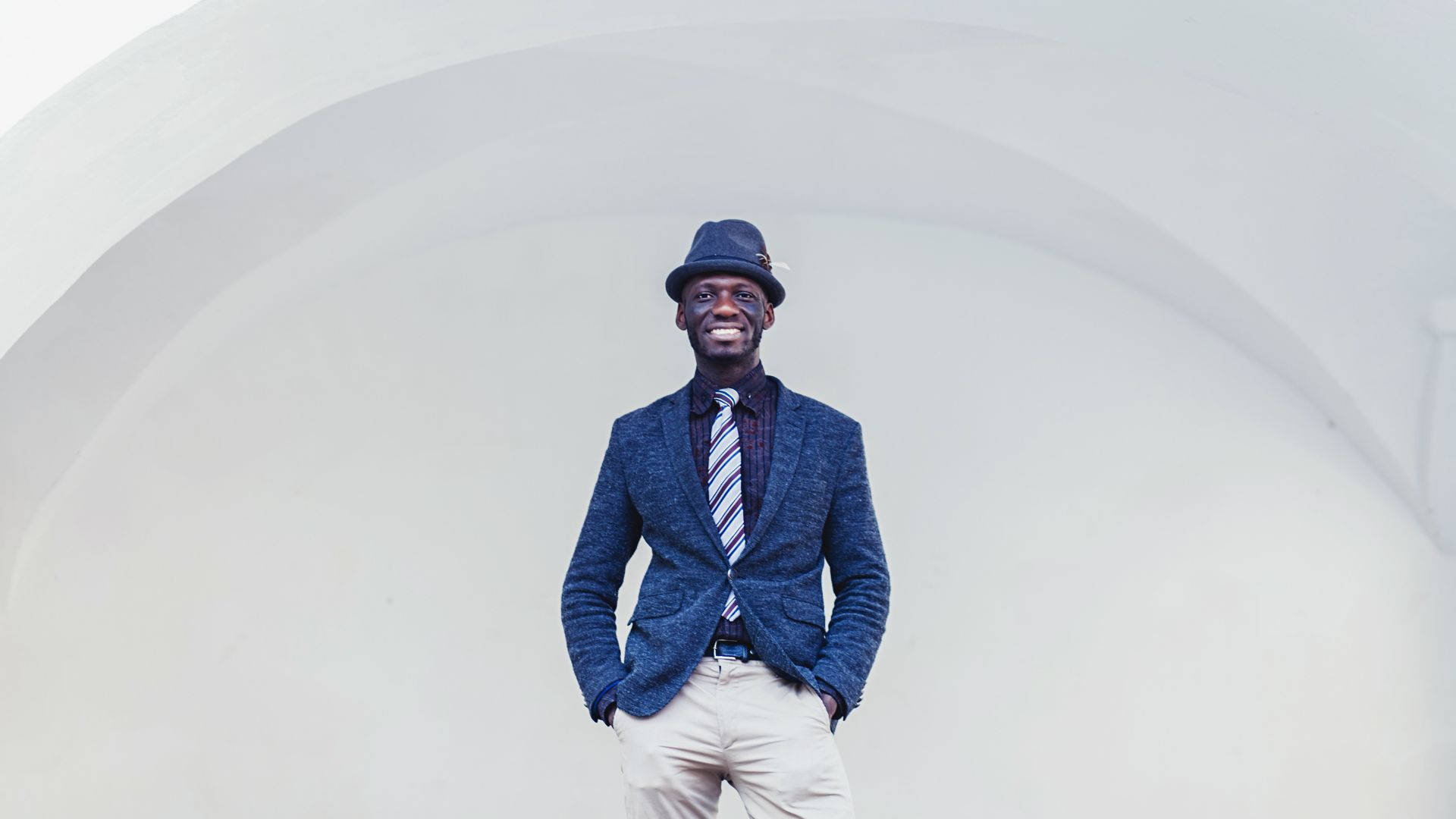 suit 桌布 壁紙 — 一位微笑的男子身穿藍色西裝外套和軟呢帽，自信地站在白色弧形建築背景前。