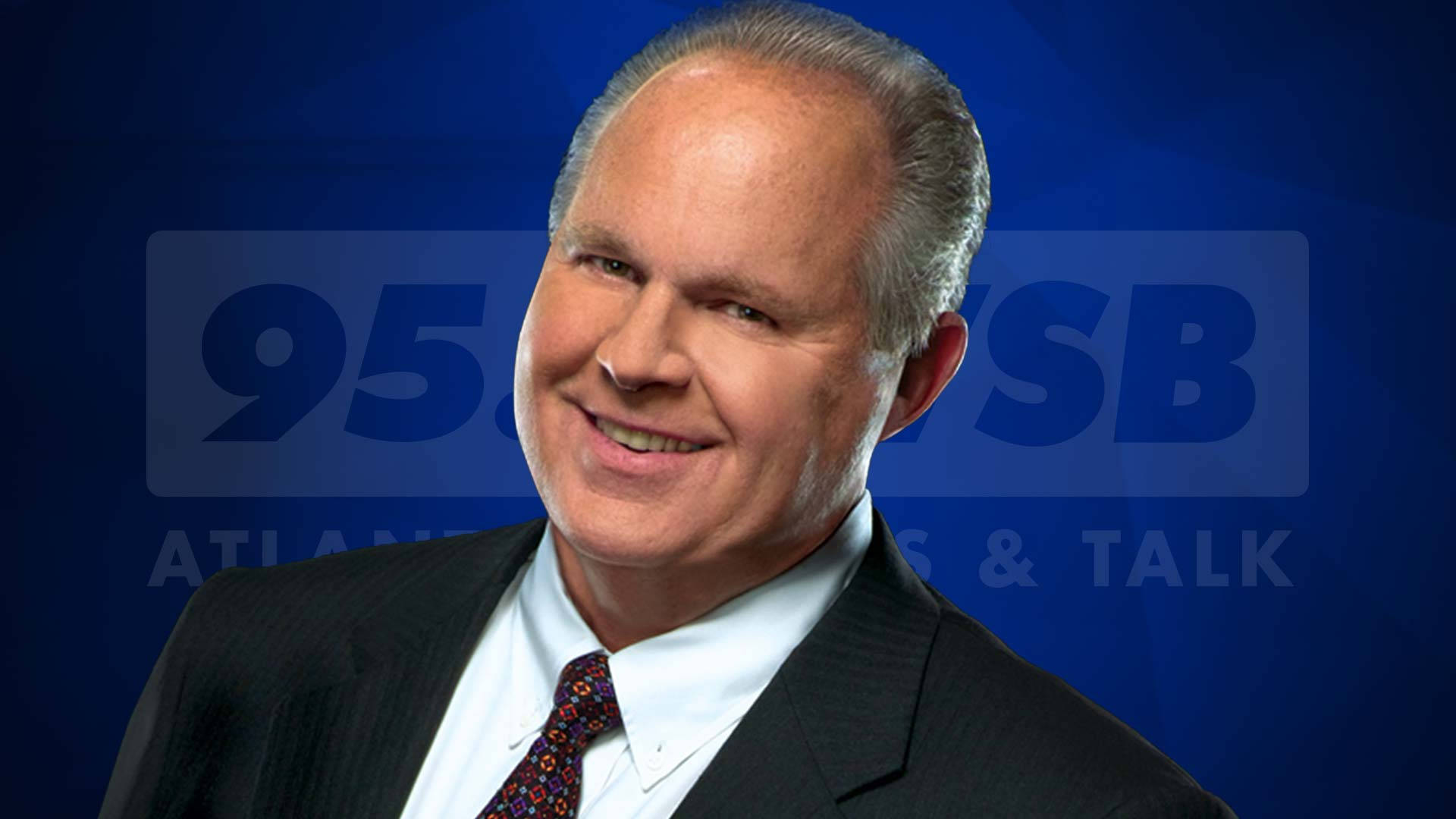 Rush 桌布 壁紙 — 一位微笑的男子身穿西裝和圖案領帶，站在帶有'95.5 WSB'品牌的藍色背景前。