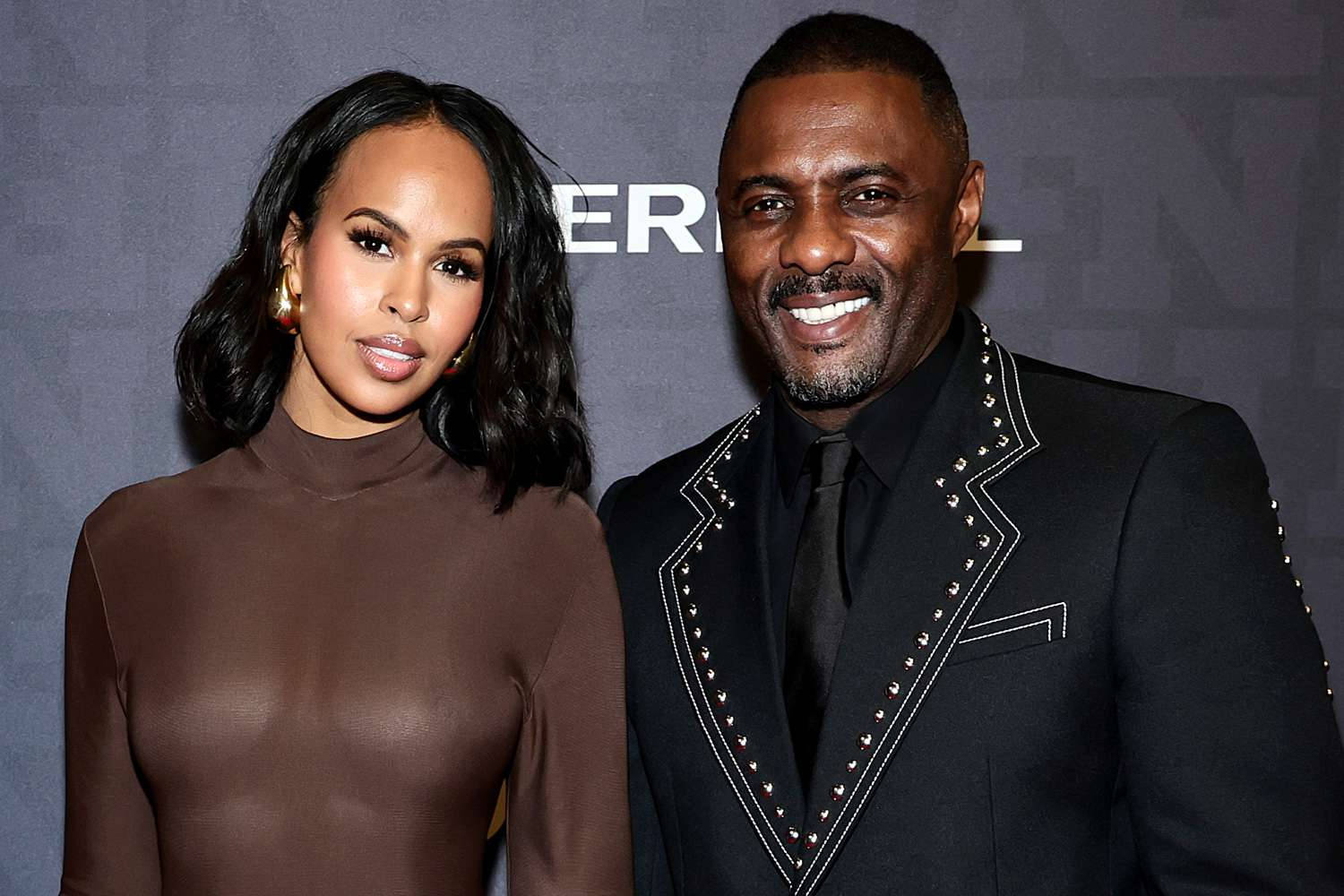idris elba 桌布 壁紙 — 一位微笑的男子身穿鑲釘黑色西裝，站在一位穿著棕色高領毛衣的女子旁邊，出席正式活動。