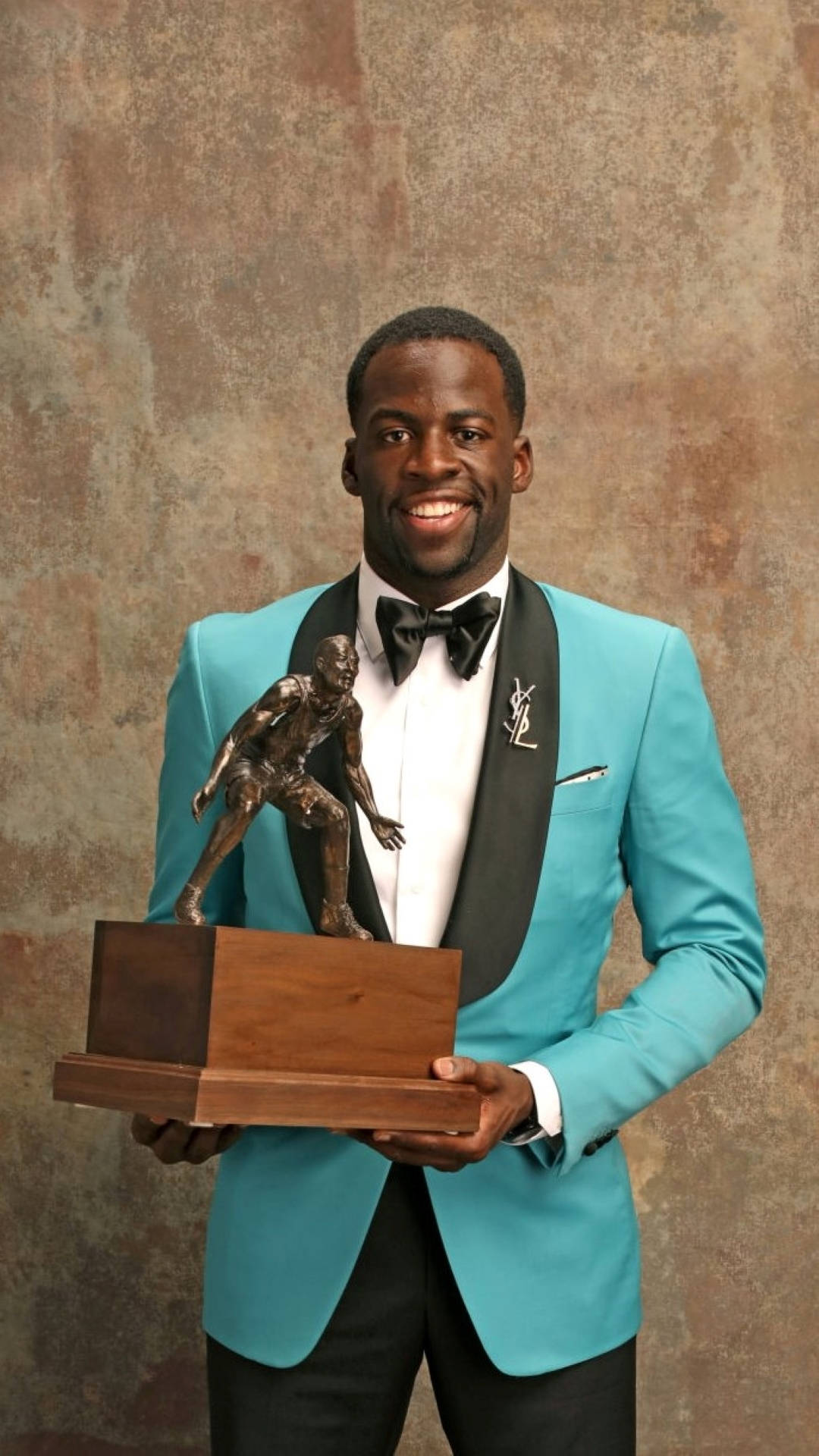 Draymond Green 桌布 壁紙 — 一位微笑的男子身穿青綠色燕尾服，在紋理棕色背景前手持足球員雕像獎盃。