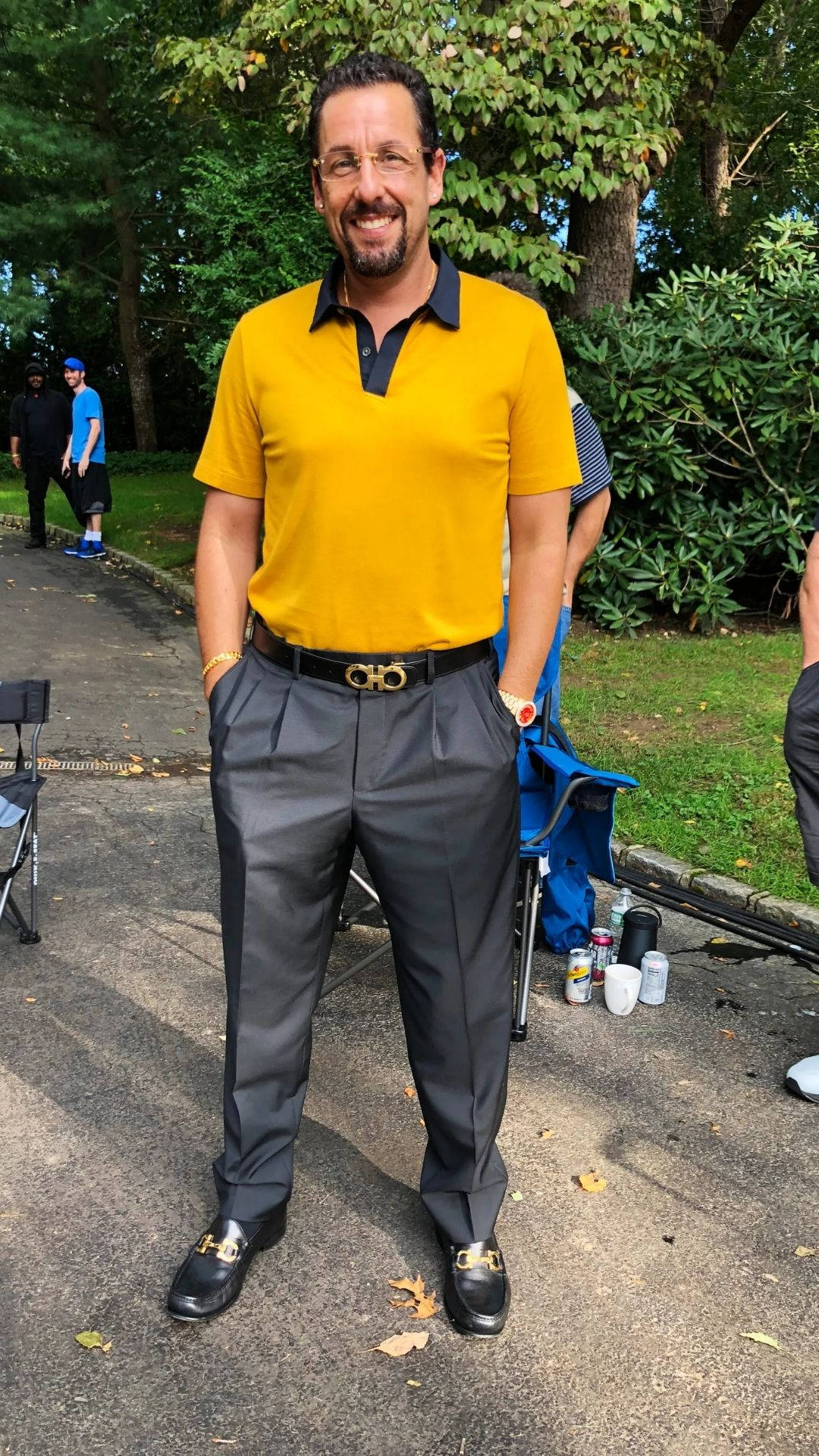 Adam Sandler 桌布 壁紙 — 一位微笑的男子身穿黃色Polo衫和灰色長褲，自信地站在戶外鋪砌的小徑上，周圍環繞著茂密的綠樹和樹葉。