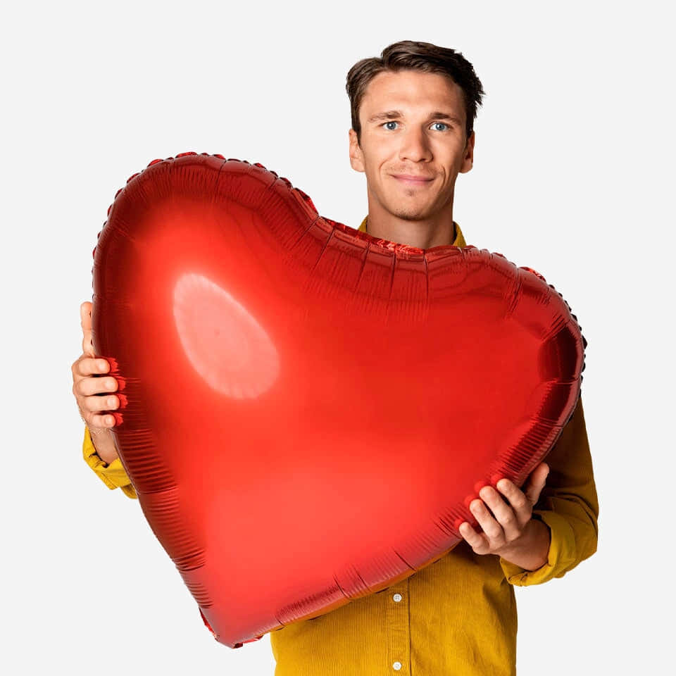 Valentine's Day 桌布 壁紙 — 一位微笑的男子身穿黃色襯衫，在純白色背景前拿著一個閃亮的大紅色心形氣球。