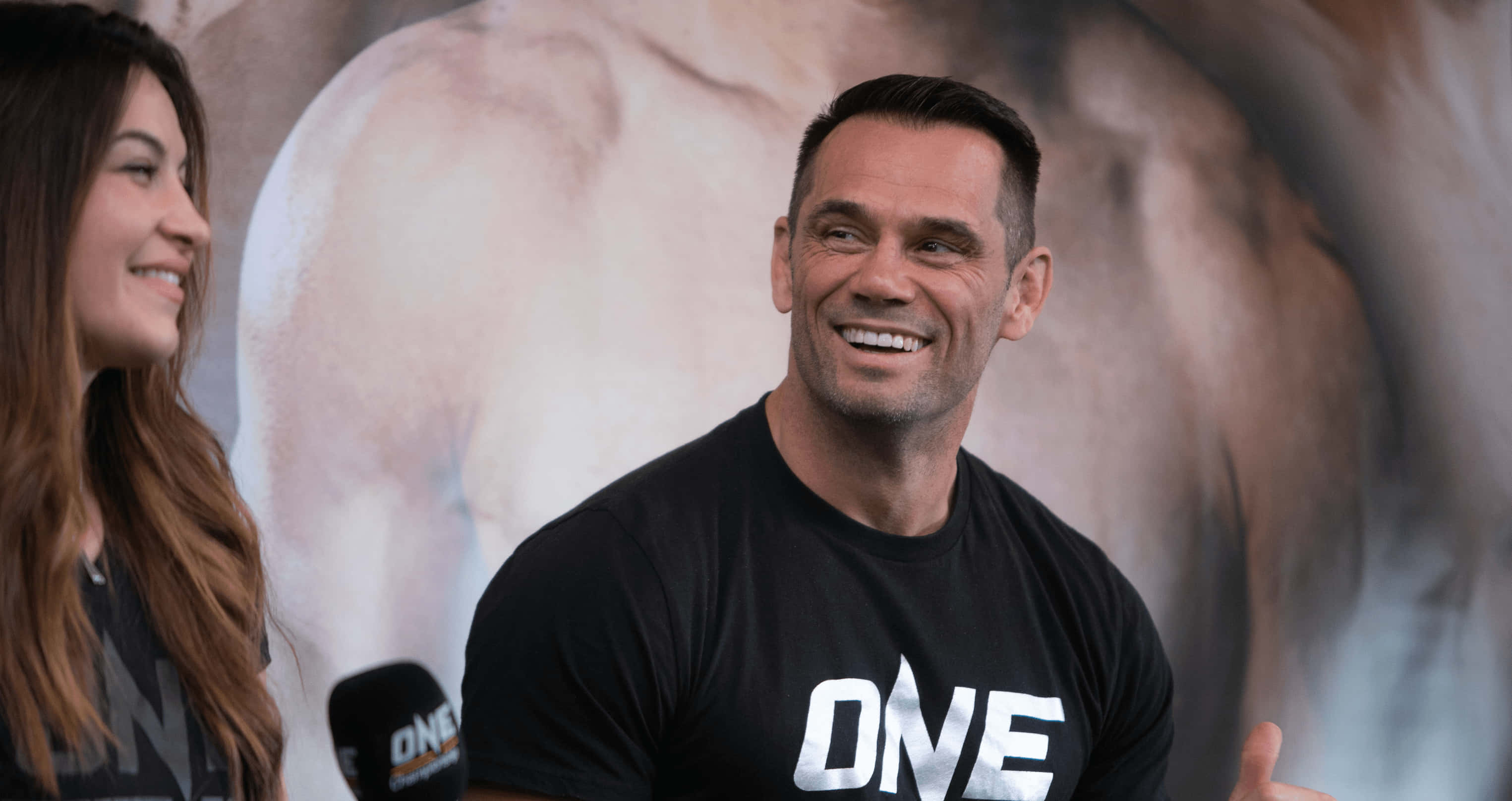 UFC 桌布 壁紙 — 一位微笑的男子身穿黑色「ONE」T恤，在記者會上向身旁的女子說話並做手勢。