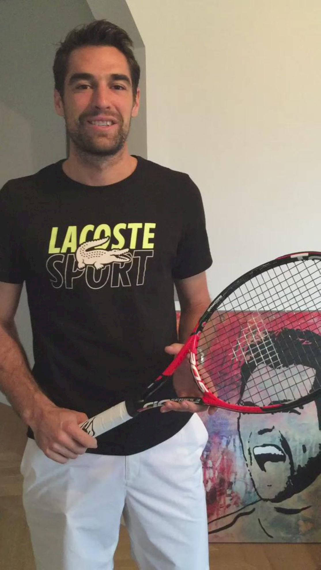 Tennis 桌布 壁紙 — 一位微笑的男子身穿黑色Lacoste Sport T恤和白色長褲，在室內手持紅色網球拍，旁邊是一幅色彩繽紛的肖像畫。