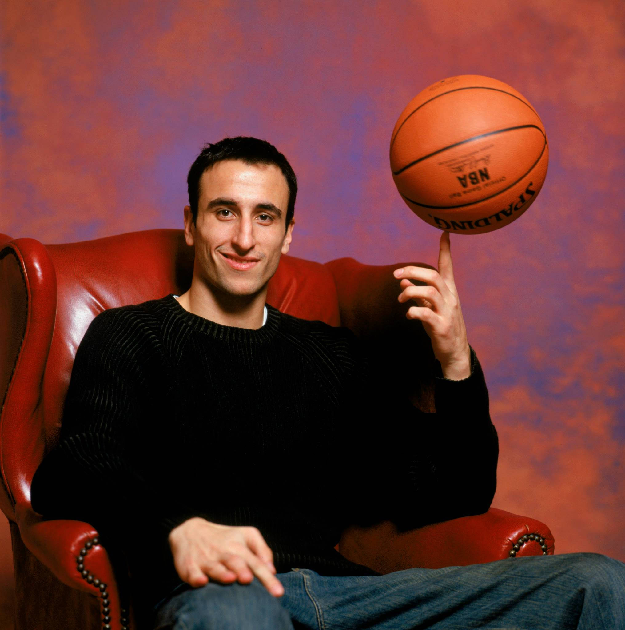 Manu 桌布 壁紙 — 一位微笑的男子身穿黑色毛衣，坐在紅色皮革扶手椅上，指尖上平衡著一個籃球，背景是帶有紋理的紅色和紫色。