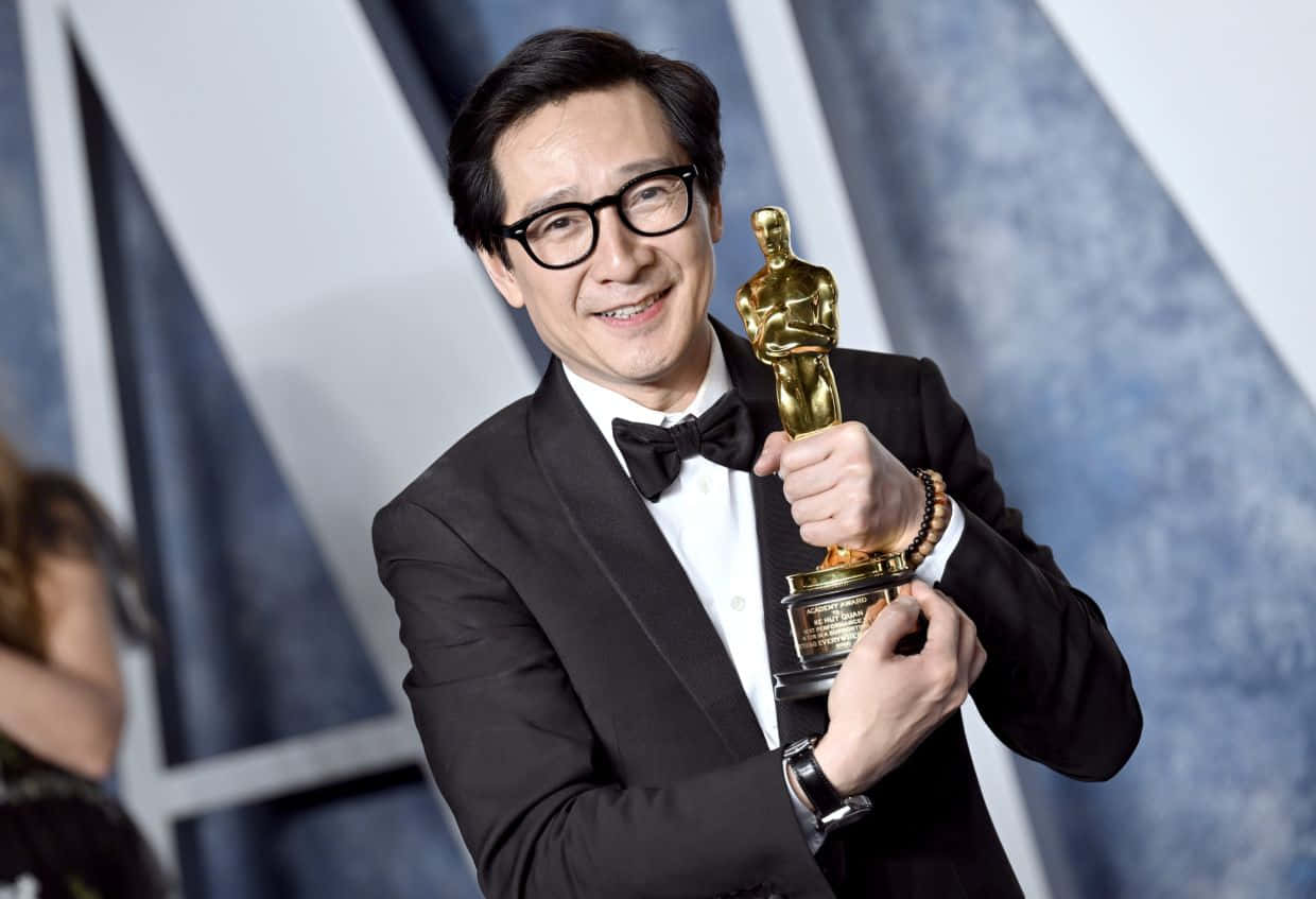 Ke Huy Quan 桌布 壁紙 — 一位微笑的男子身穿黑色燕尾服、戴著眼鏡，在幾何藍白背景前自豪地手持金色奧斯卡獎座。