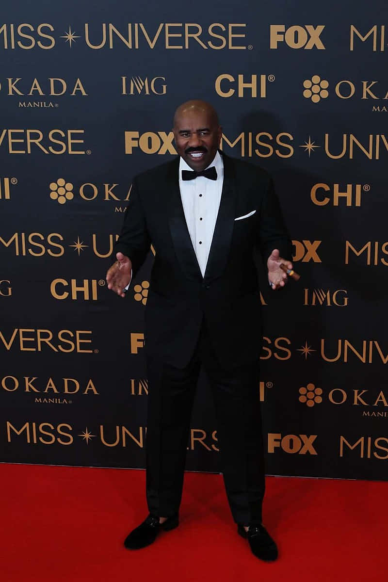 steve harvey 桌布 壁紙 — 一位微笑的男子身穿黑色燕尾服配蝴蝶結，站在品牌背景前的紅地毯上。