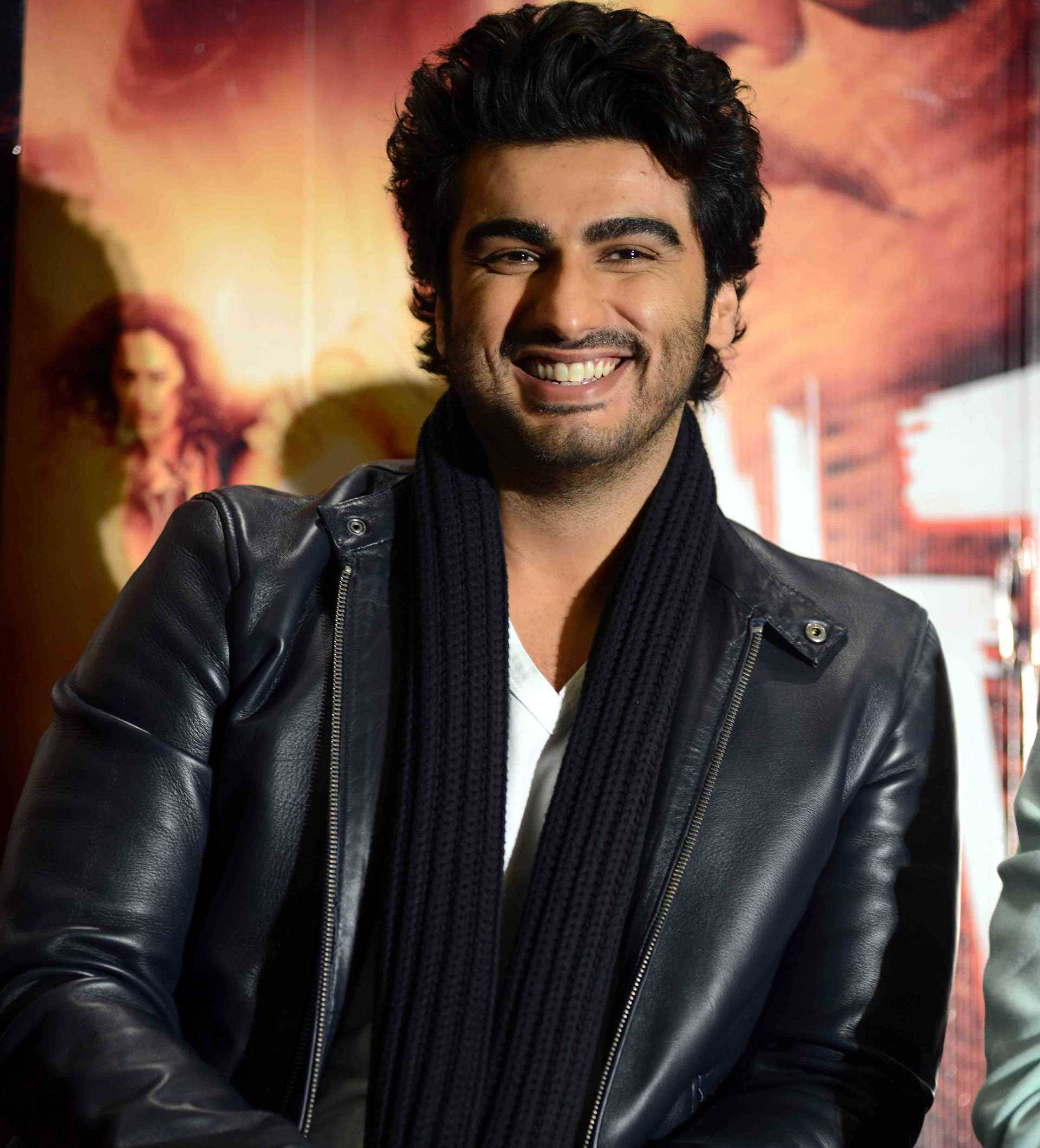 Arjun Kapoor 桌布 壁紙 — 一位微笑的男子身穿黑色皮夾克和圍巾，在暖色調的電影海報背景前自信地擺姿勢。
