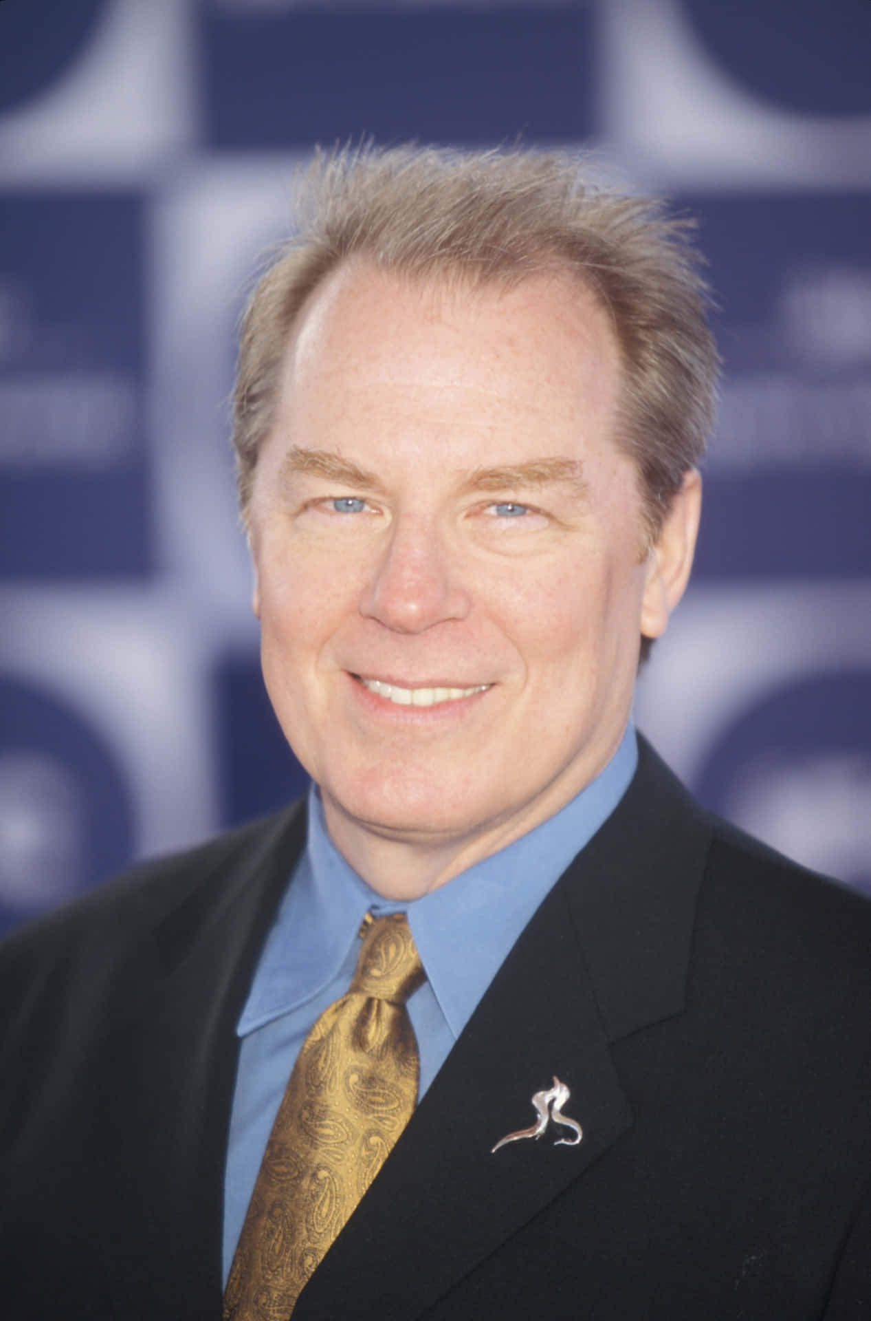 Michael Mckean 桌布 壁紙 — 一位微笑的男子身穿黑色西裝、藍色襯衫和金色佩斯利領帶，在模糊的藍白格子背景前擺姿勢。