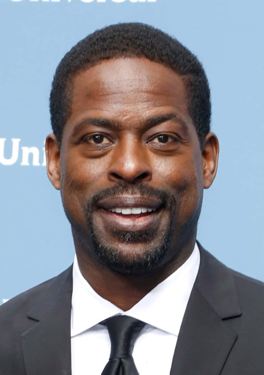 Sterling K Brown 桌布 壁紙 — 一位微笑的男子身穿黑色西裝及領帶，在淺藍色背景前擺姿勢，背景有部分文字可見。