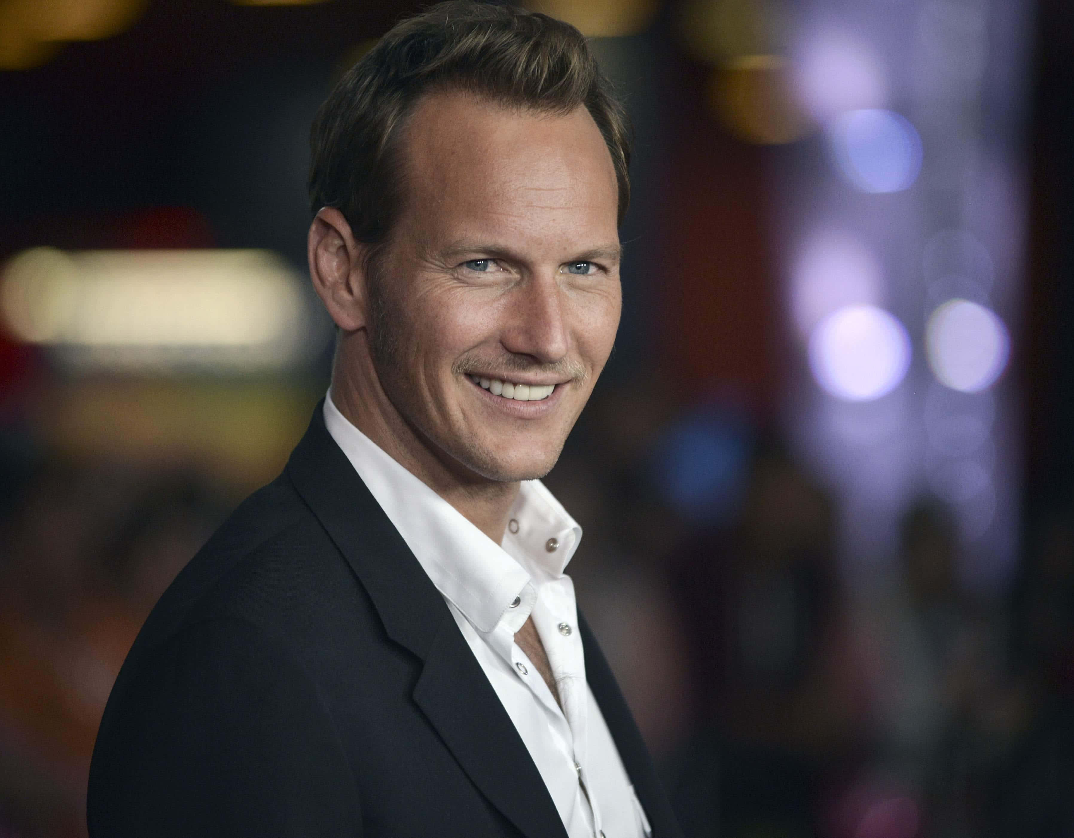 Patrick Wilson 桌布 壁紙 — 一位微笑的男子身穿黑色西裝和白色襯衫，站在色彩繽紛的散景燈光模糊背景前。