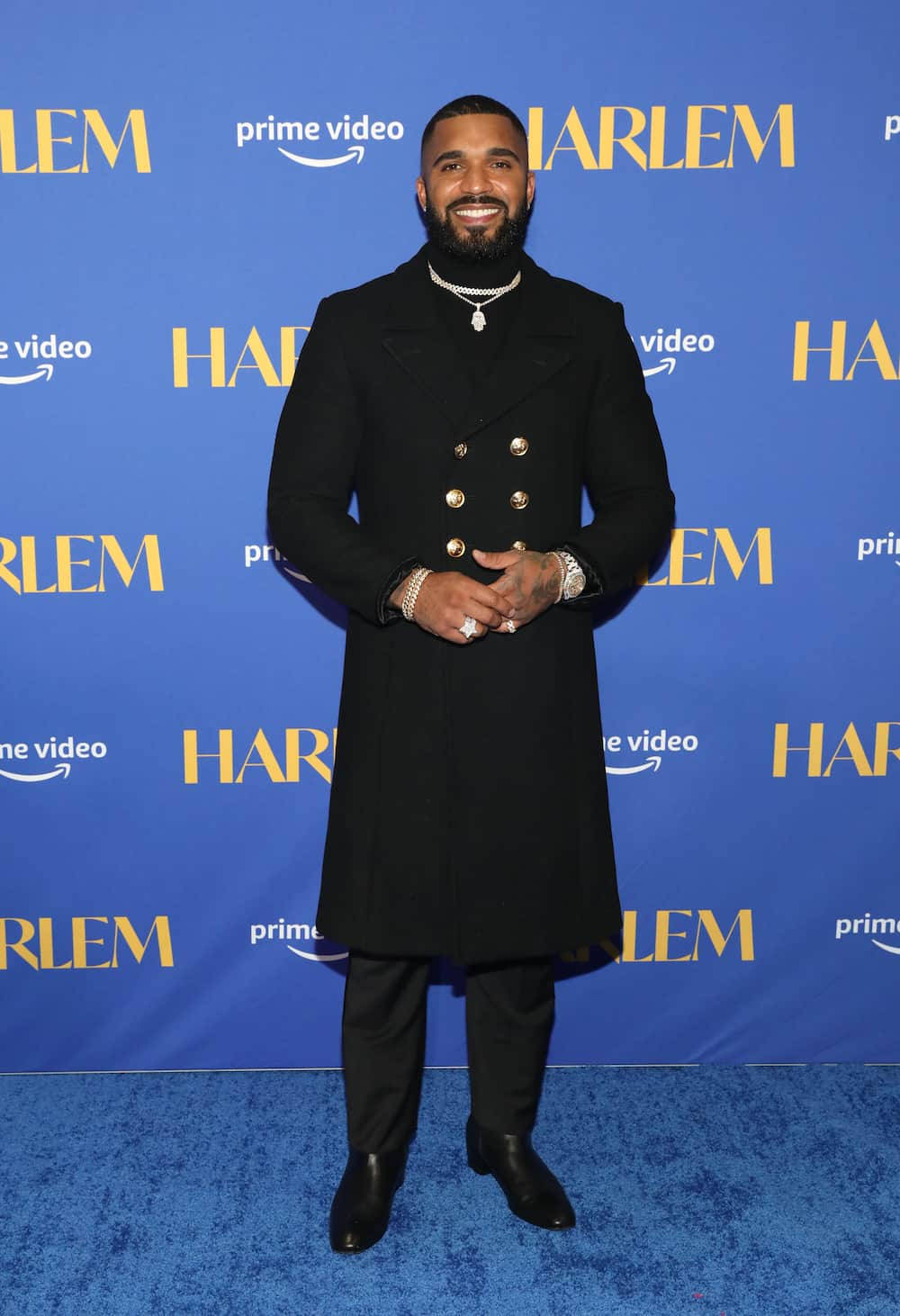 Black Man 桌布 壁紙 — 一位微笑的男子身穿黑色雙排扣大衣，自信地站在藍色地毯上，背景為品牌標誌。