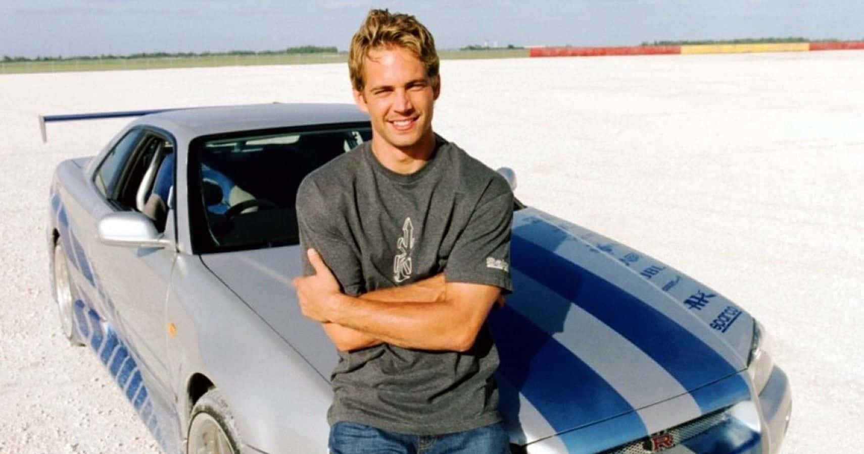 Paul Walker Skyline 桌布 壁紙 — 一位微笑的男子雙臂交叉，自信地站在一輛銀藍色跑車旁，背景是廣闊的白色鹽灘，陽光普照。
