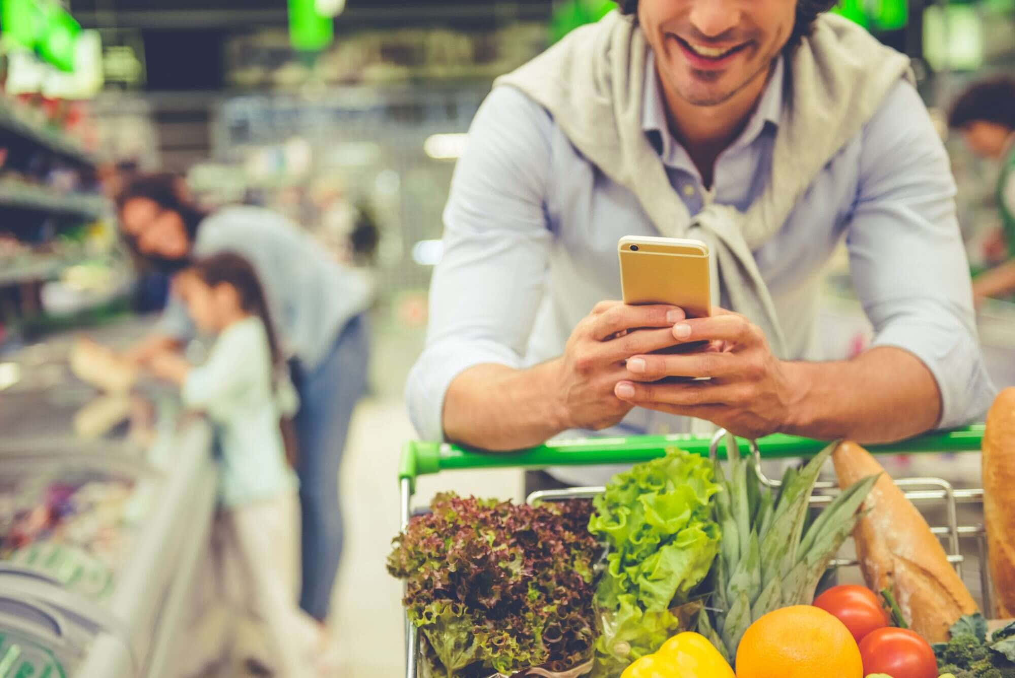 Grocery Store 桌布 壁紙 — 一位微笑的男子靠在裝滿新鮮農產品的購物車上，同時在光線明亮的超市裡查看手機。