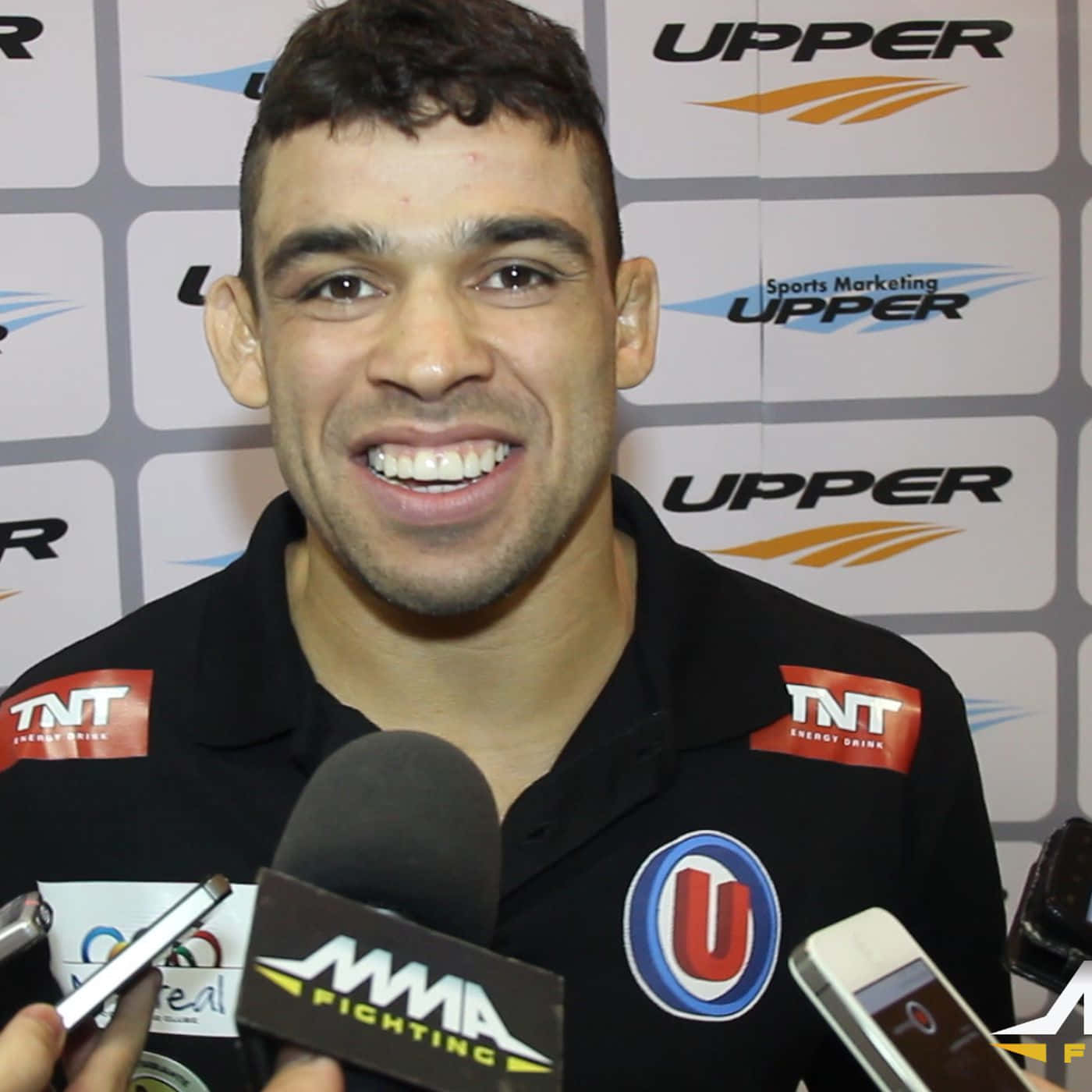 UFC 桌布 壁紙 — 一位微笑的男性拳手穿著黑色品牌Polo衫，在記者會上向記者發言，身後有麥克風和贊助商標誌。