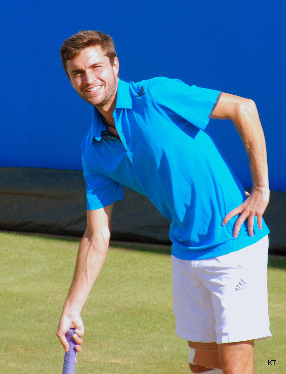 Tennis 桌布 壁紙 — 一位微笑的男性網球運動員穿著亮藍色Polo衫和白色短褲，在草地場上靠著鮮豔的藍色牆壁隨意倚靠。