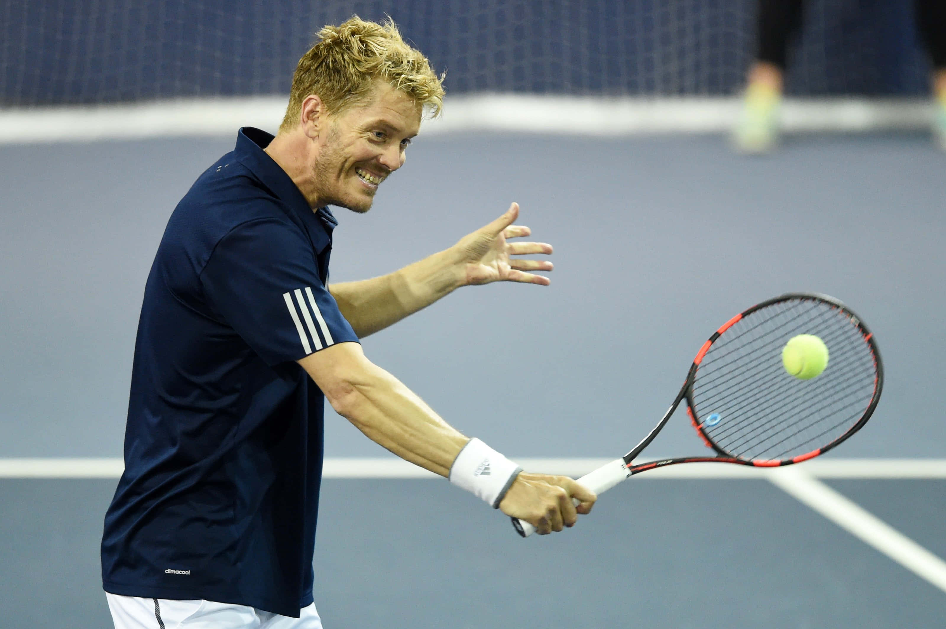 Tennis 桌布 壁紙 — 一位微笑的男性網球運動員身穿海軍藍襯衫，在藍色球場上揮拍擊打黃色網球。