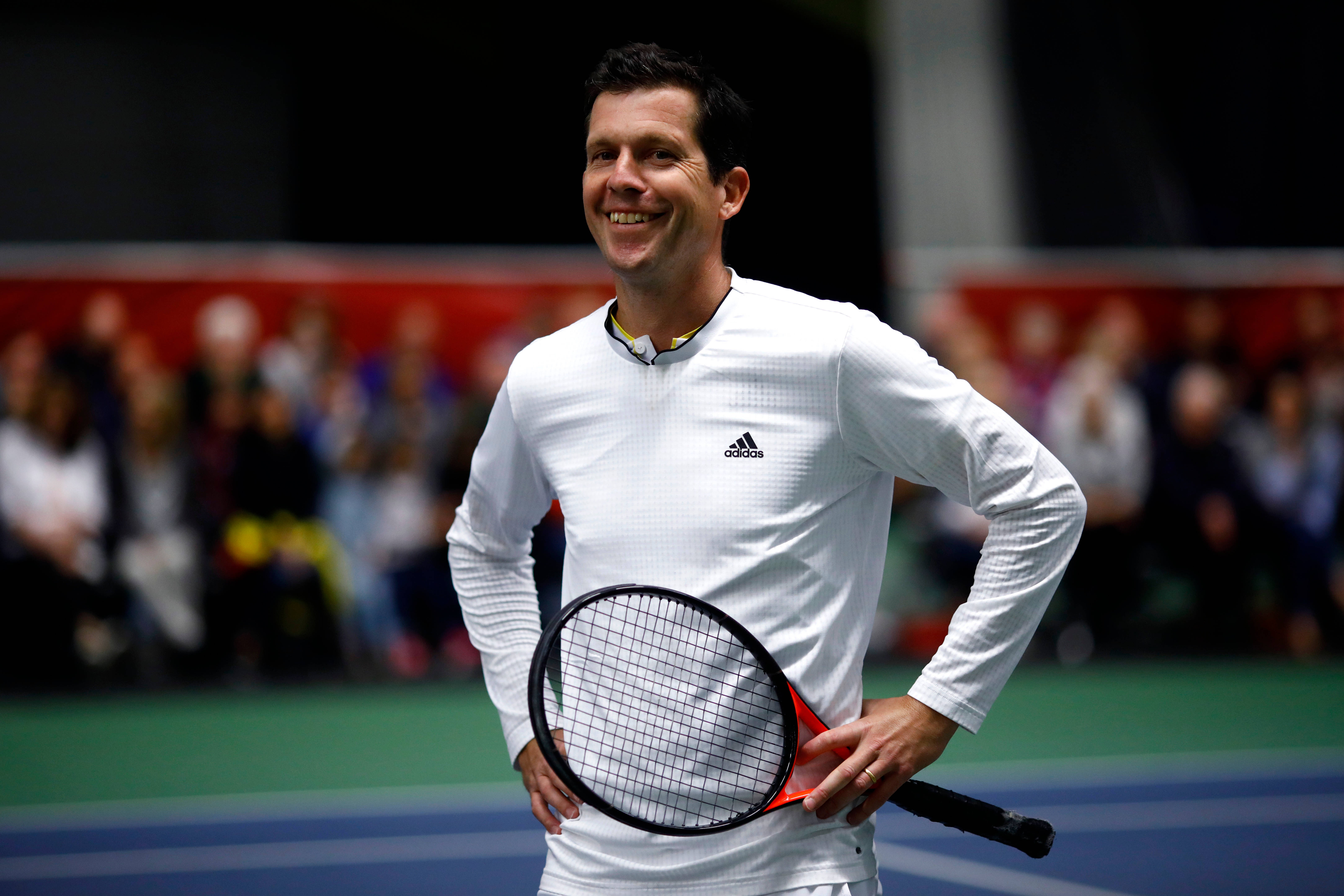 網球 桌布 壁紙 — 一位微笑的男性網球選手穿著白色Adidas裝備，在充滿活力的藍綠色球場上自信地握著球拍，身後有模糊的觀眾。