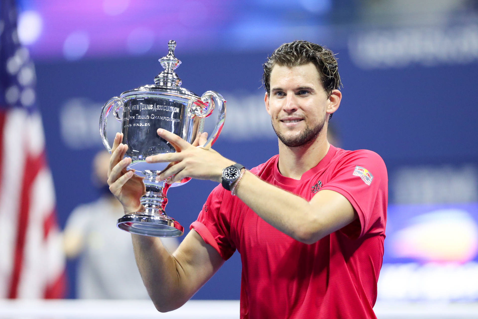 dominic thiem 桌布 壁紙 — 一位微笑的男性網球選手穿著紅色襯衫，在可見美國國旗的球場上自豪地舉起一個大銀色獎盃。