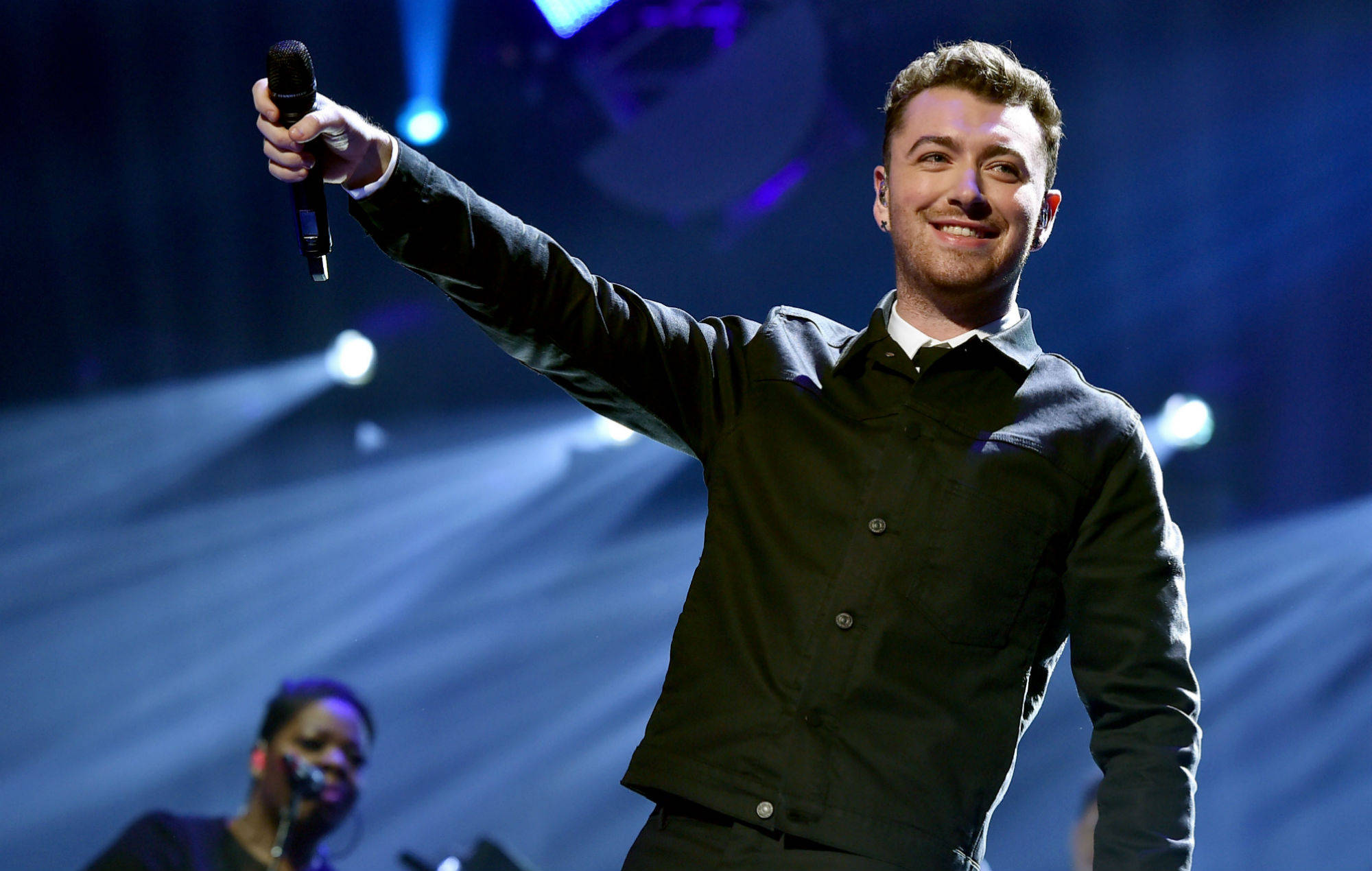 Sam Smith 桌布 壁紙 — 一位微笑的男歌手身穿深色外套，在明亮的舞台燈光下高舉麥克風，光束穿過藍色煙霧。