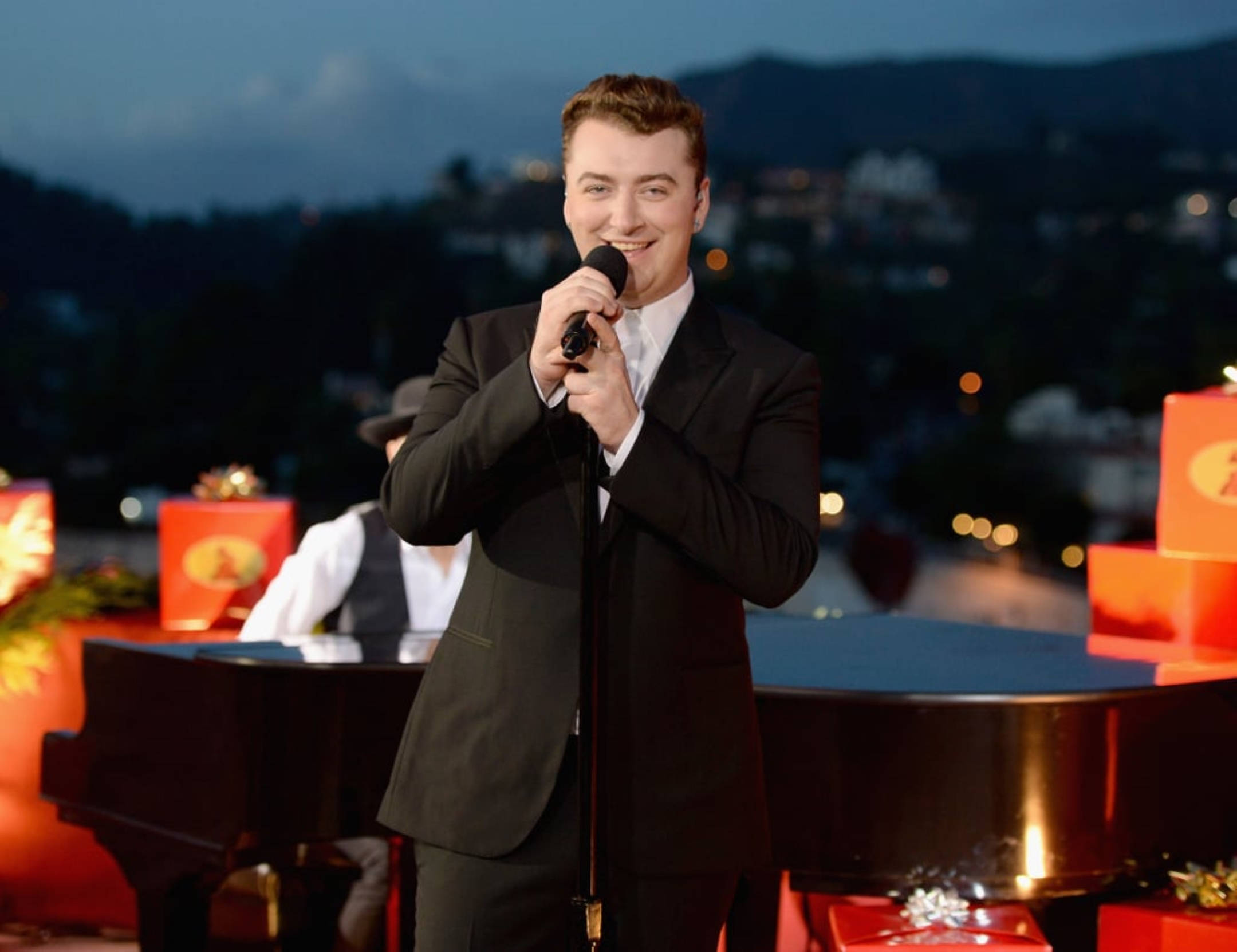 Sam Smith 桌布 壁紙 — 一位微笑的男歌手身穿黑色西裝，在黃昏時分於屋頂舞台上表演，背景中城市燈光閃爍。