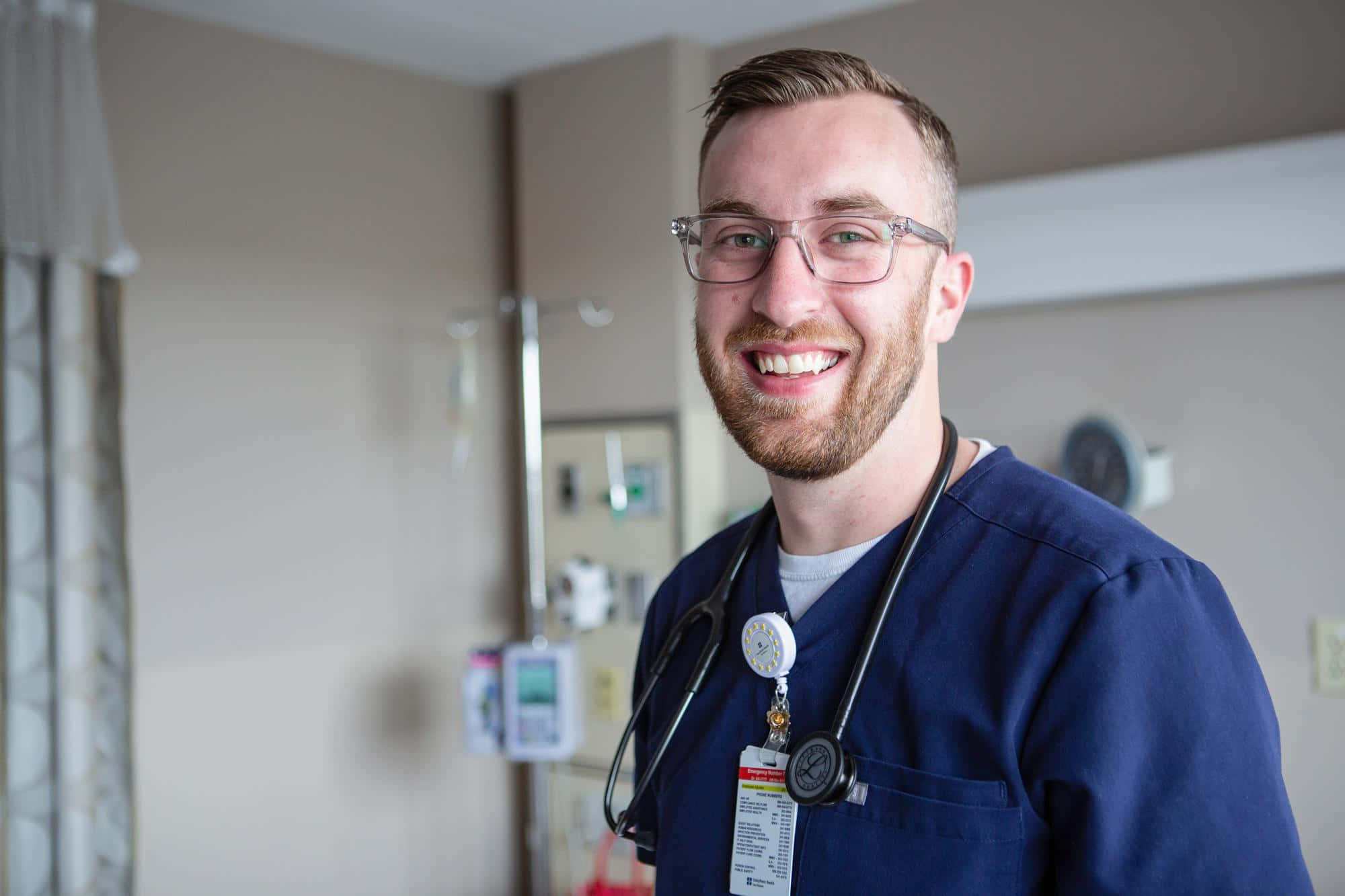 Nursing 桌布 壁紙 — 一位微笑的男護士穿著藍色手術衣和眼鏡，站在醫院房間內，身後有一個輸液架。