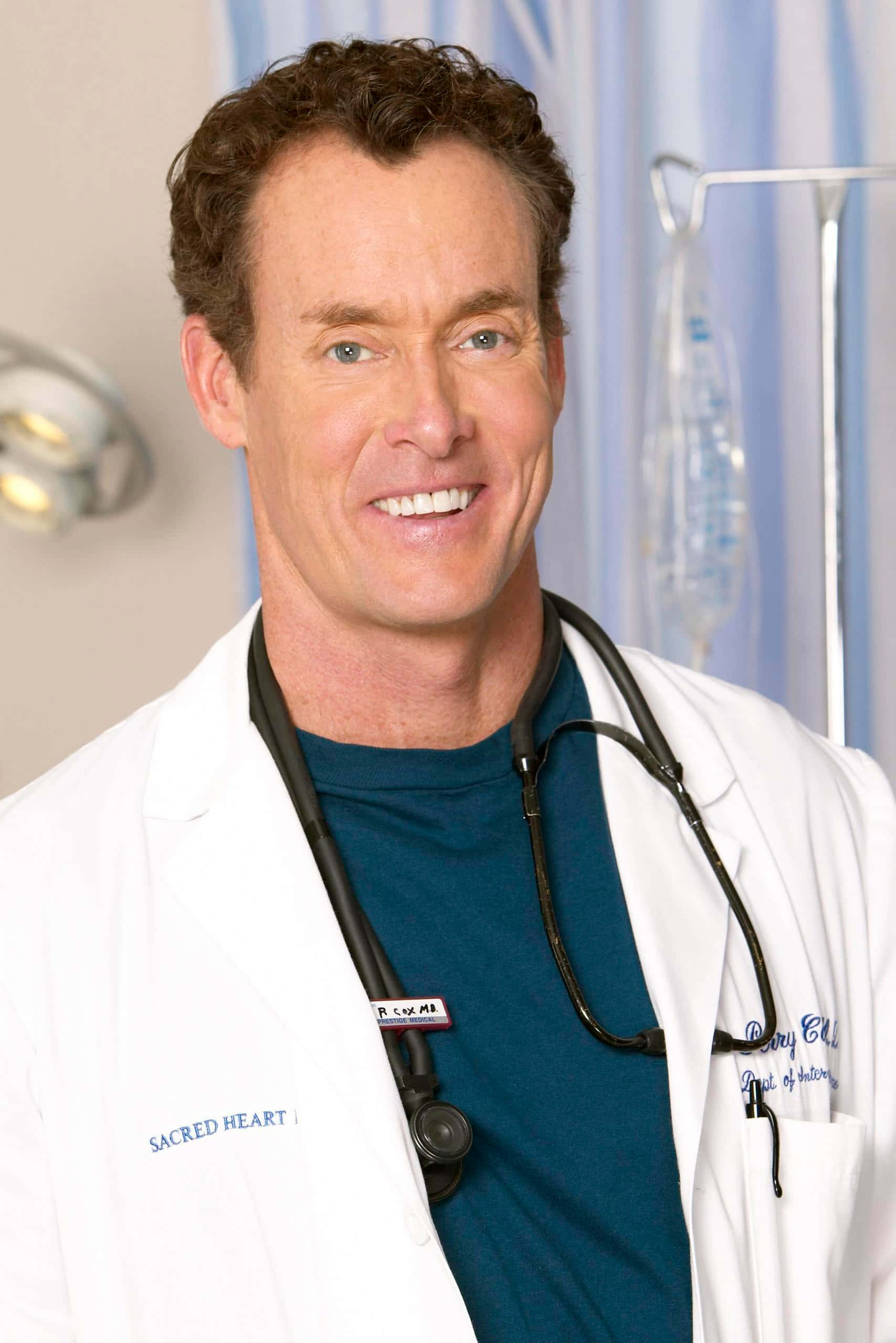 John C. Mcginley 桌布 壁紙 — 一位微笑的男醫生穿著白色實驗室外套，掛著聽診器，站在一間身後有醫療設備的醫院房間裡。